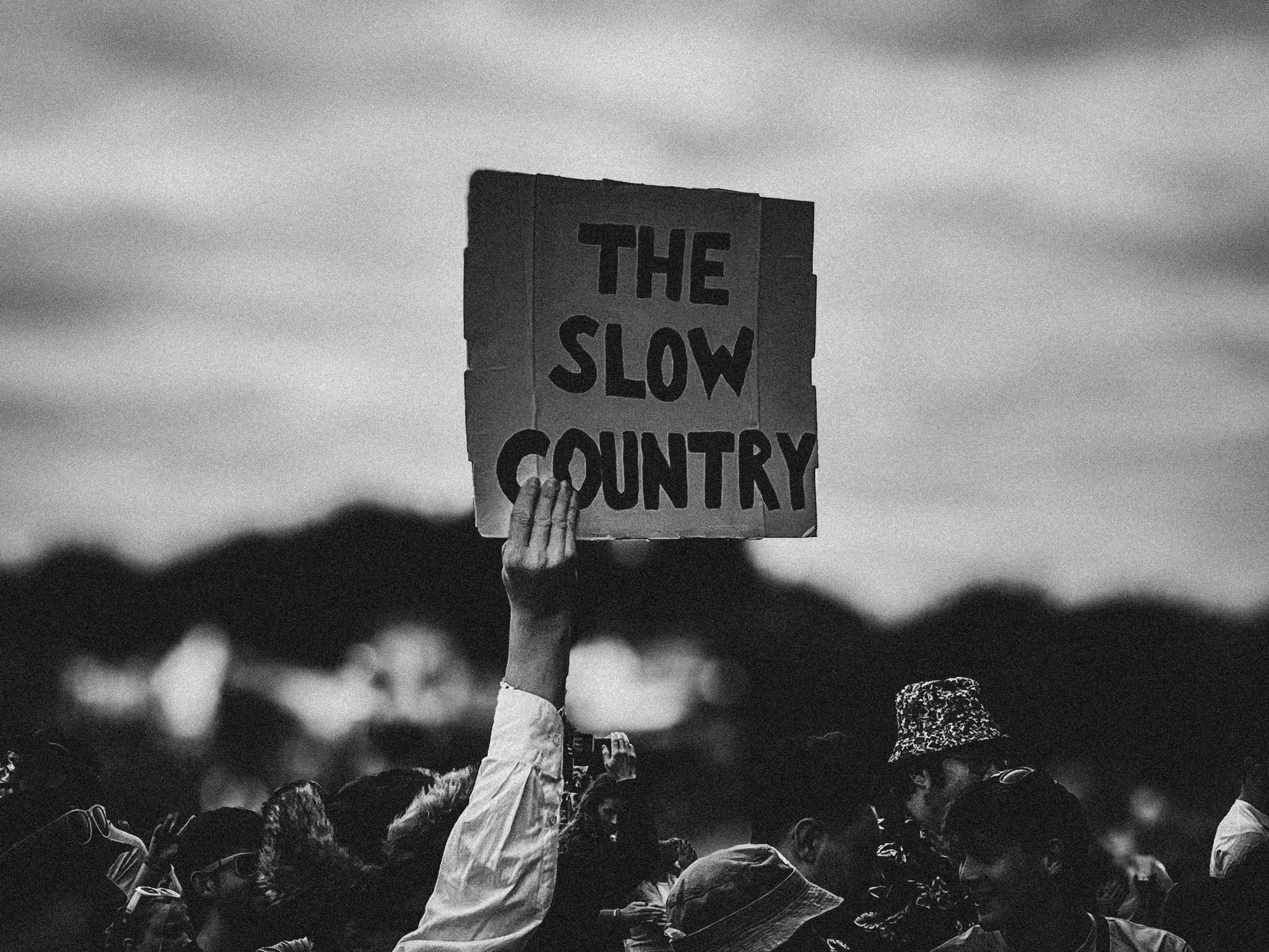 A black and white photo of a crowd at a protest or rally with a person holding a sign that reads 'THE SLOW COUNTRY'.