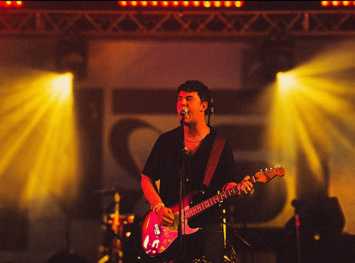 A young man playing an electric guitar and singing into a microphone on stage lit by yellow stage lights. Basht. truck festival 2025
