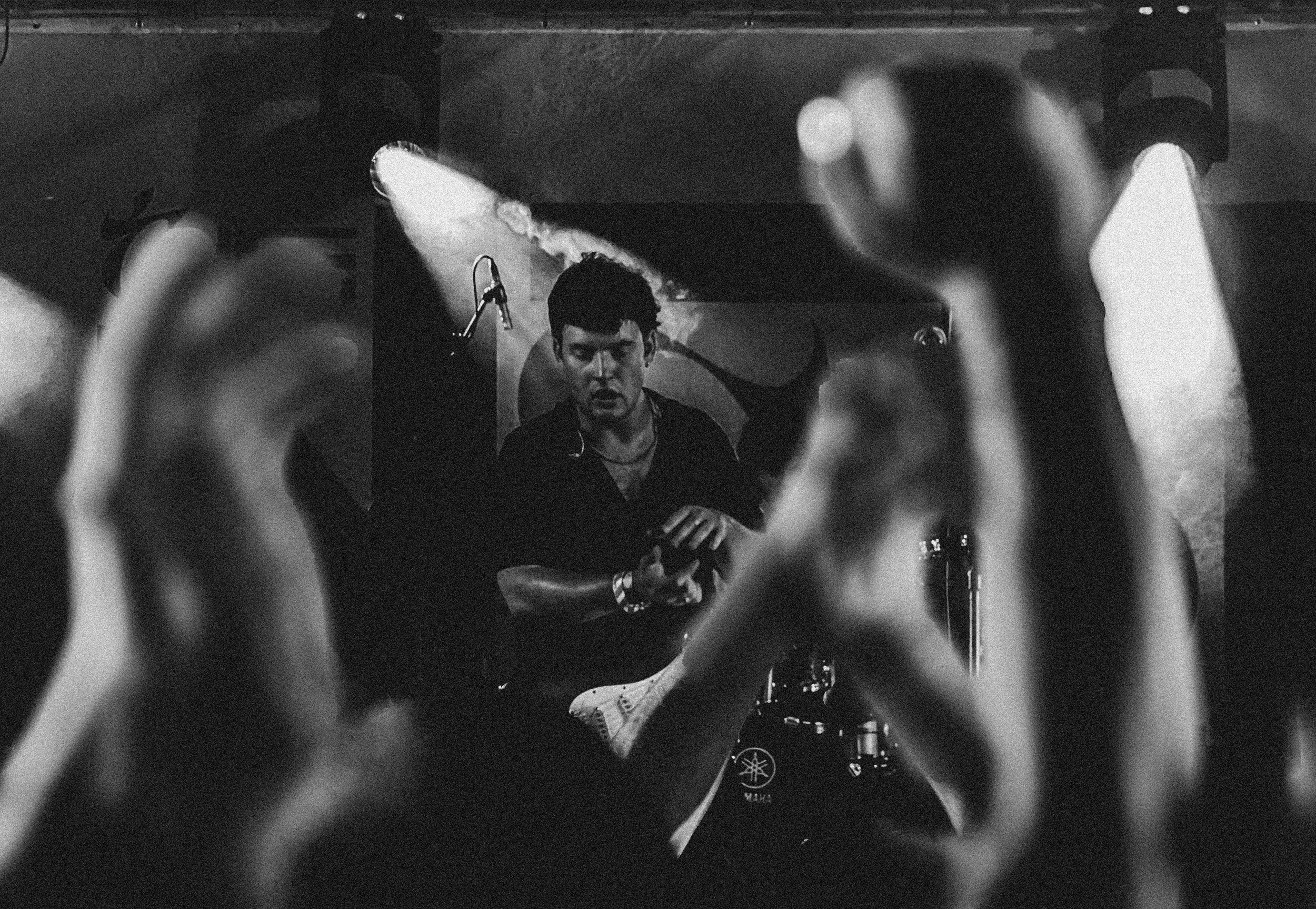 A black and white photo of a man on stage, seen through the hands of an audience member who is holding a smartphone, capturing the performance.