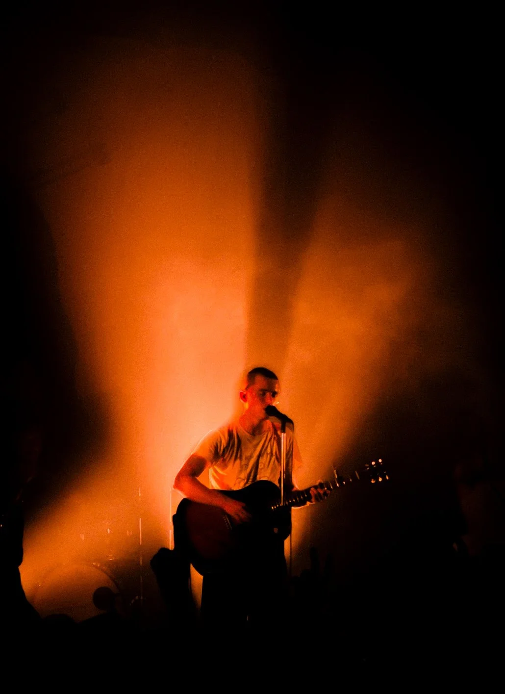 Person playing guitar and singing into a microphone on stage with warm orange lighting creating a dramatic effect. The Royston Club, Songs for the Spine Tour, Electric Bristol.