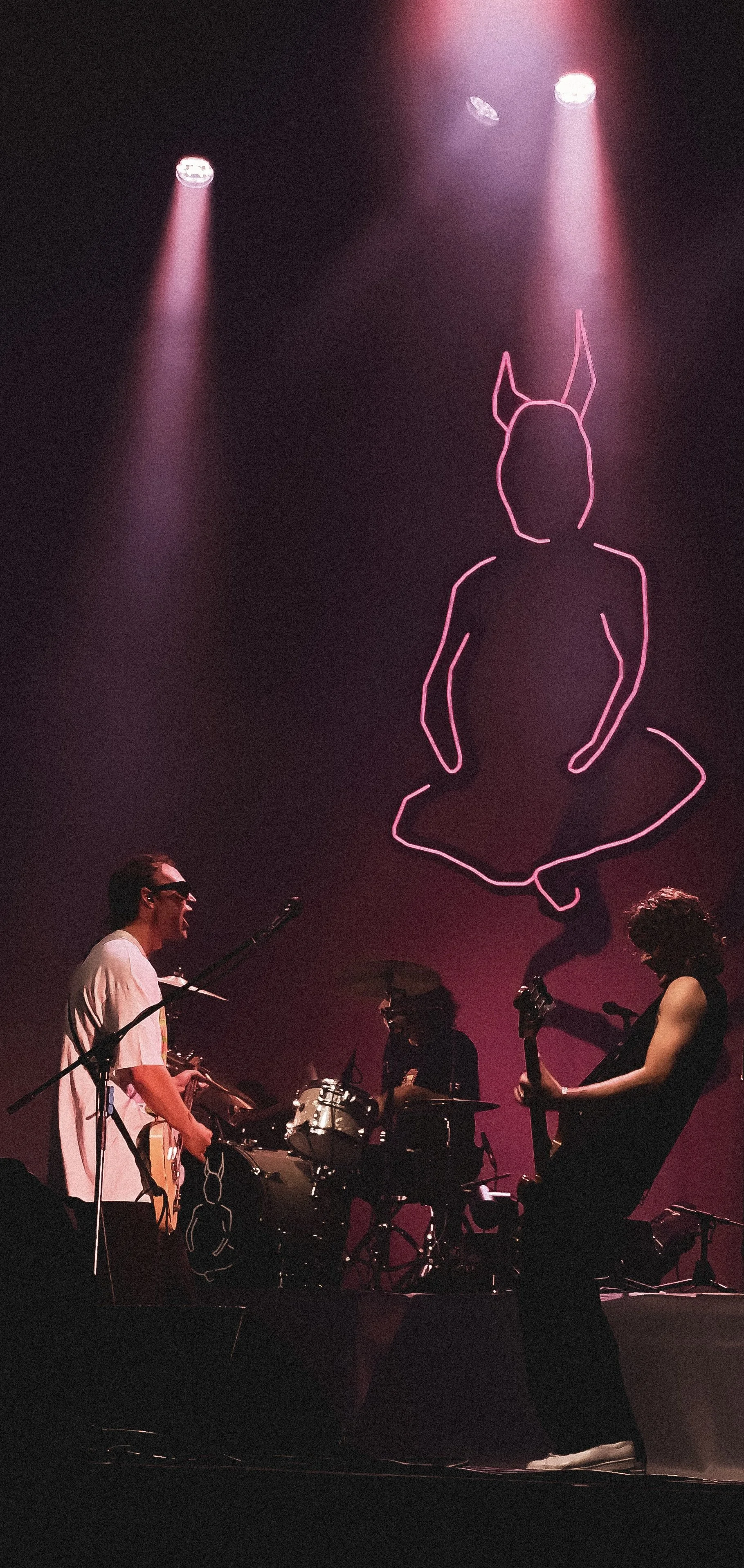 A band performing on stage with pink lighting, featuring a neon outline of a sitting person with bunny ears projected on the backdrop.