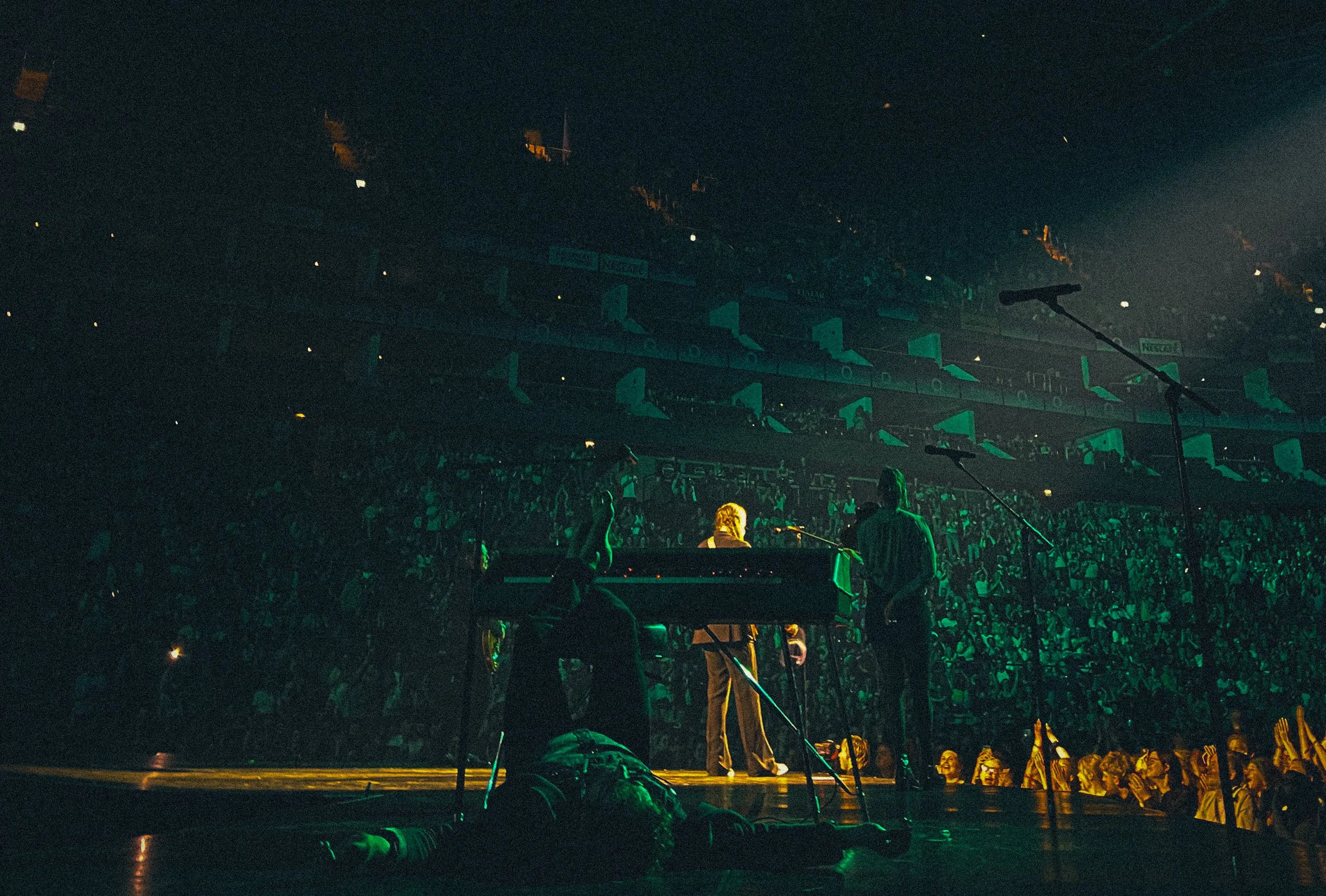 A performer on stage with three people around, in front of a large audience at an indoor concert or event, illuminated by green lighting.
