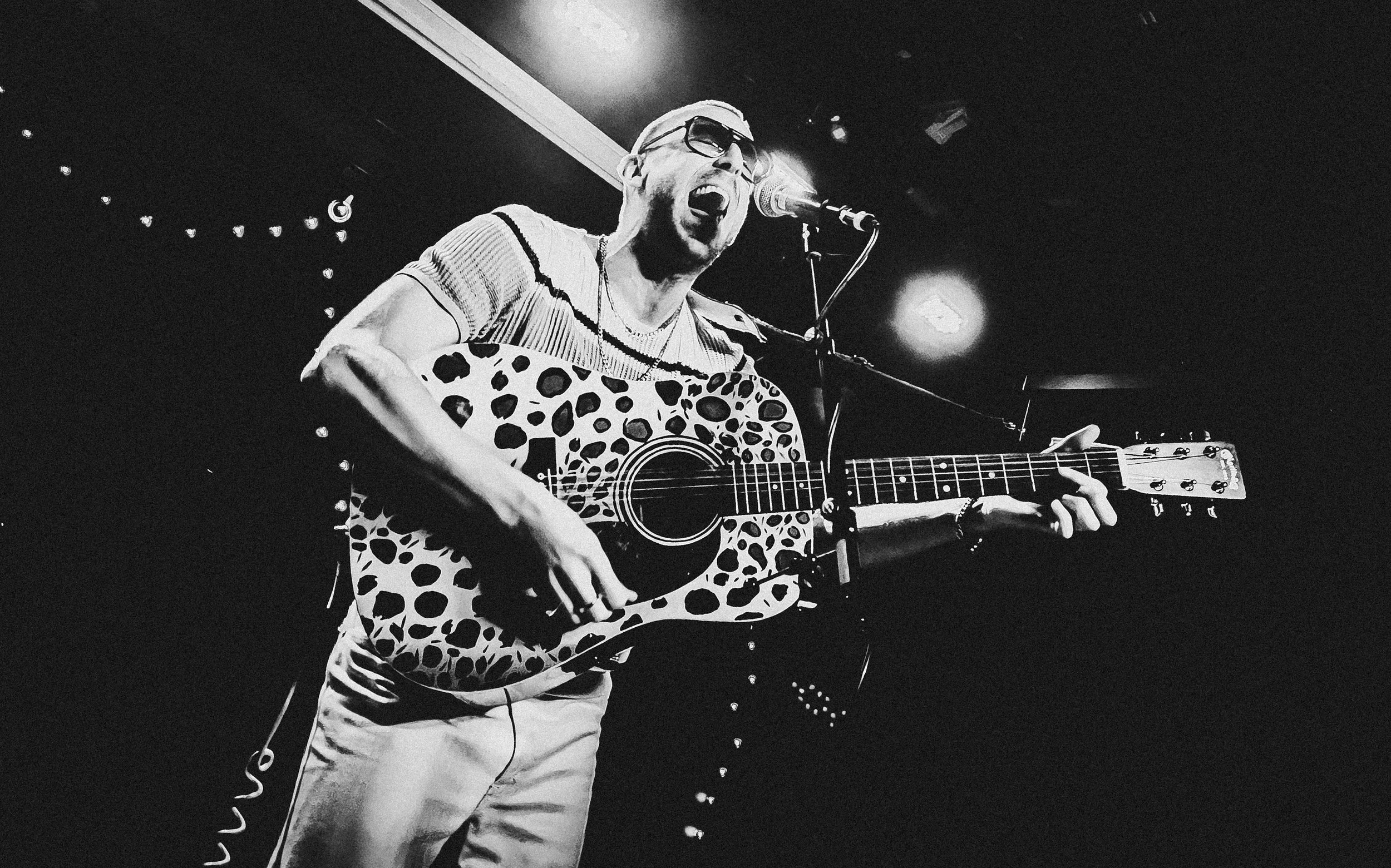 A man singing passionately while playing an acoustic guitar with a leopard print pattern on stage under dim lighting. Miles Kane acoustic sessions for Sunlight in the Shadows, Rough Trade, Bristol.