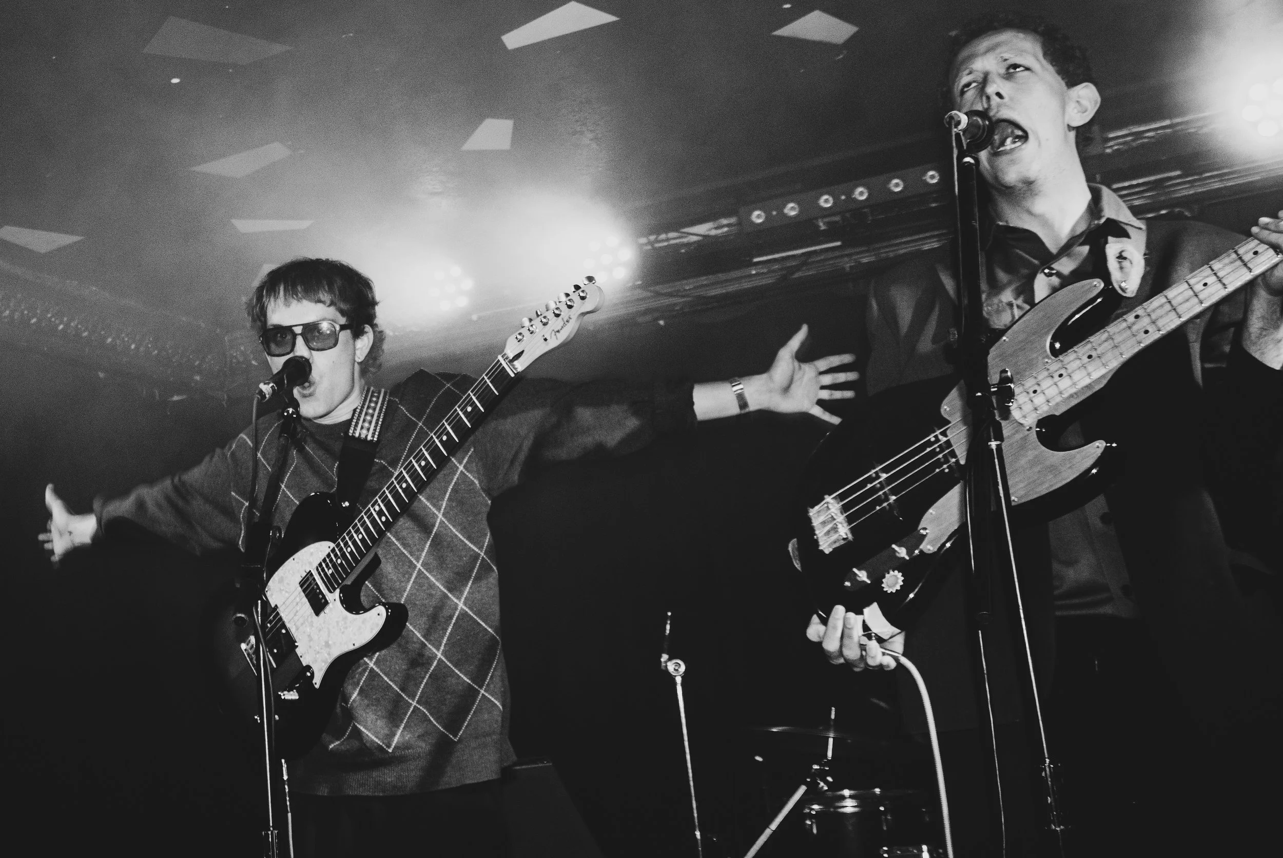 Two male musicians performing on stage with guitars and microphones, one wearing sunglasses and the other singing passionately.