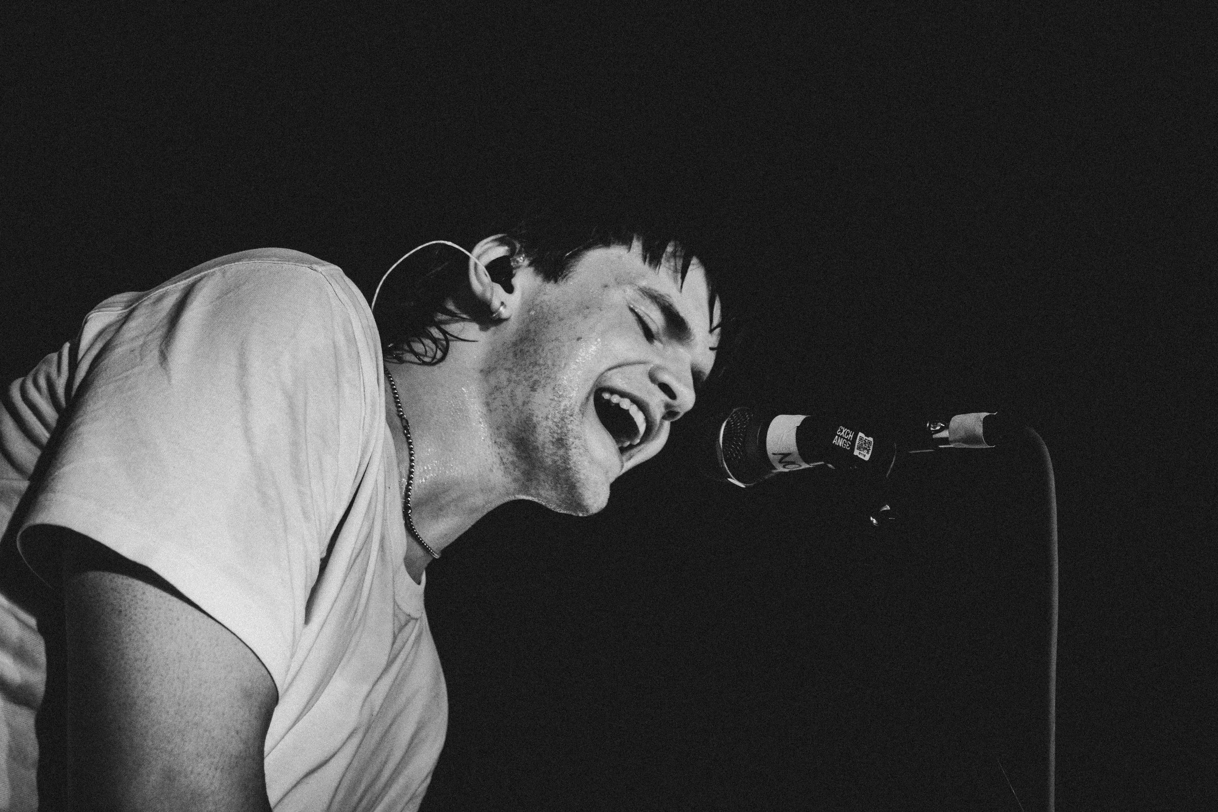 A black and white image of a man singing passionately into a microphone on stage, with his eyes closed, wearing a light-colored t-shirt and a necklace. Teenage Dads lead singer 