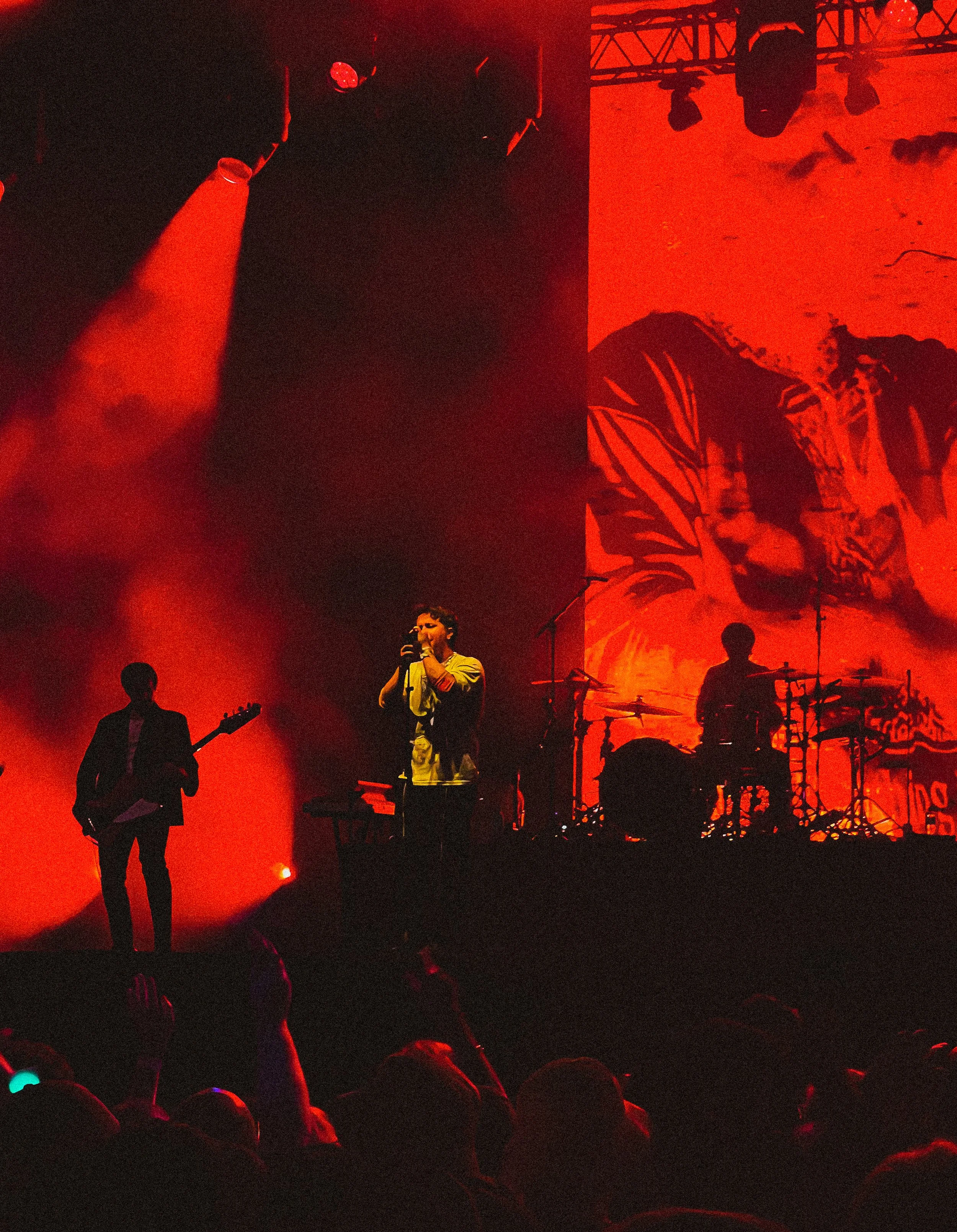 A live concert with a band on stage under red lighting, featuring a vocalist, a guitarist, and a drummer with a large red backdrop.