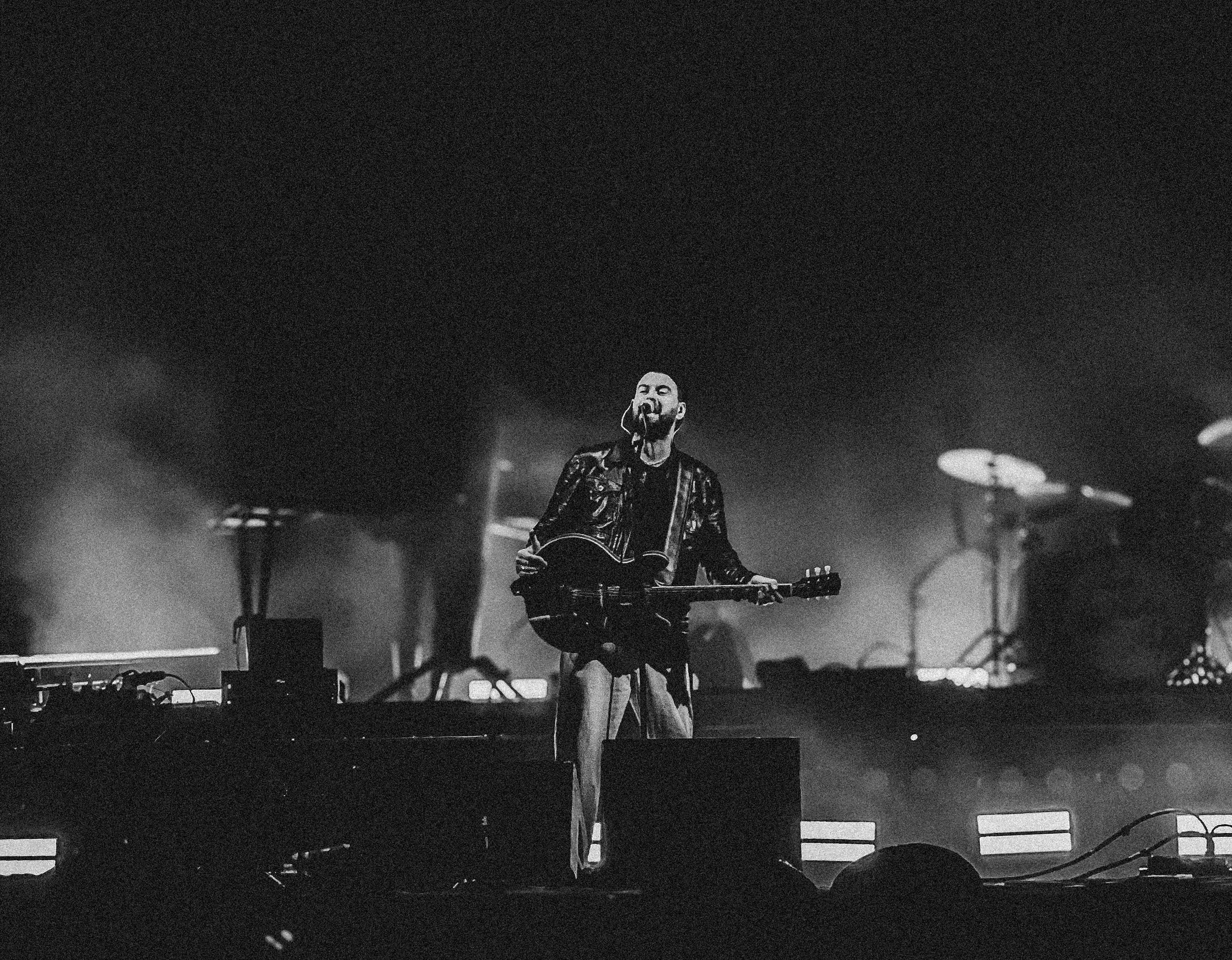 A black-and-white photo of a man singing and playing guitar on stage during a concert, with drums in the background and stage lights creating a dramatic atmosphere. The Courteeners at Truck Festival