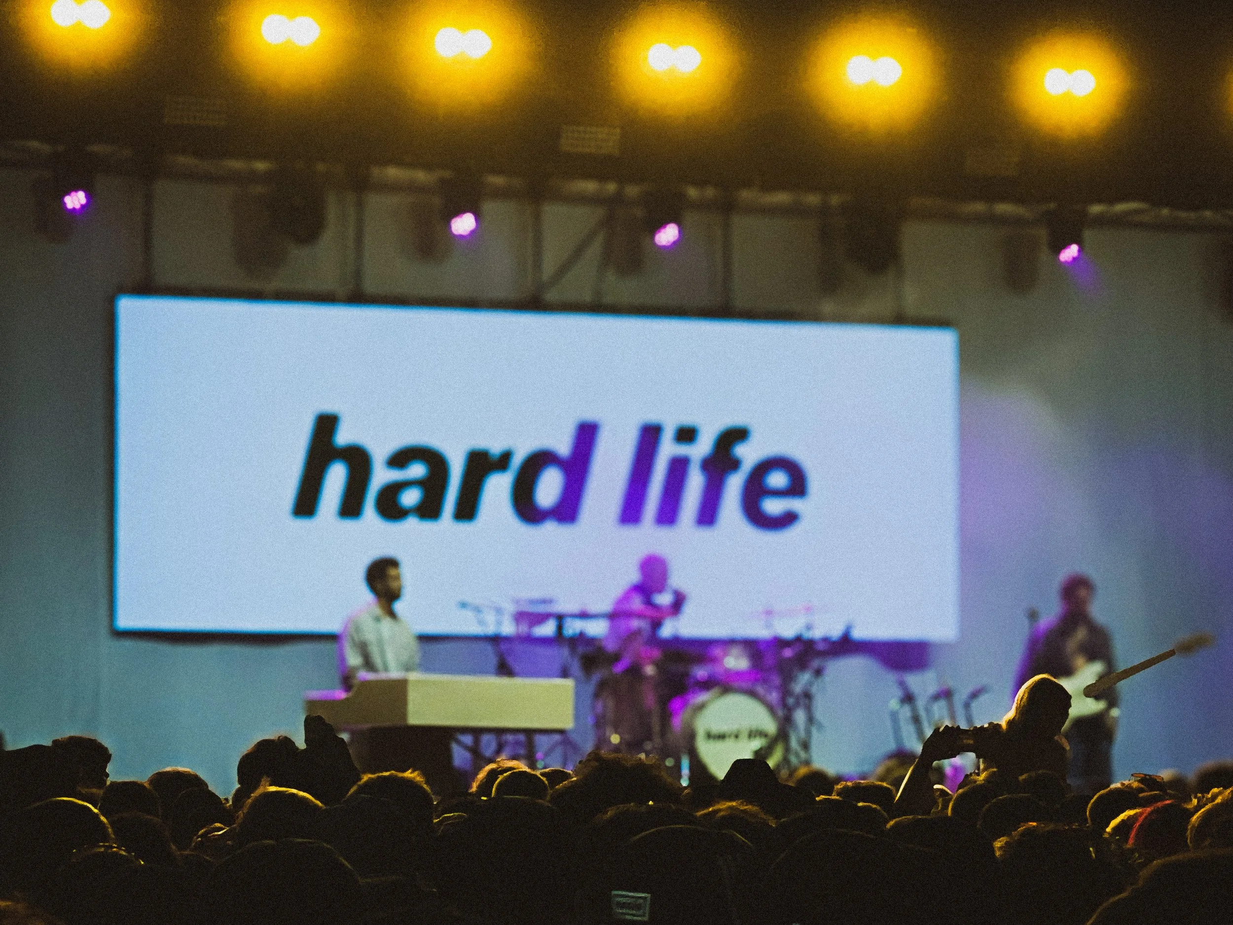 Concert stage with band playing; large screen displaying the words 'hard life'; audience watching.