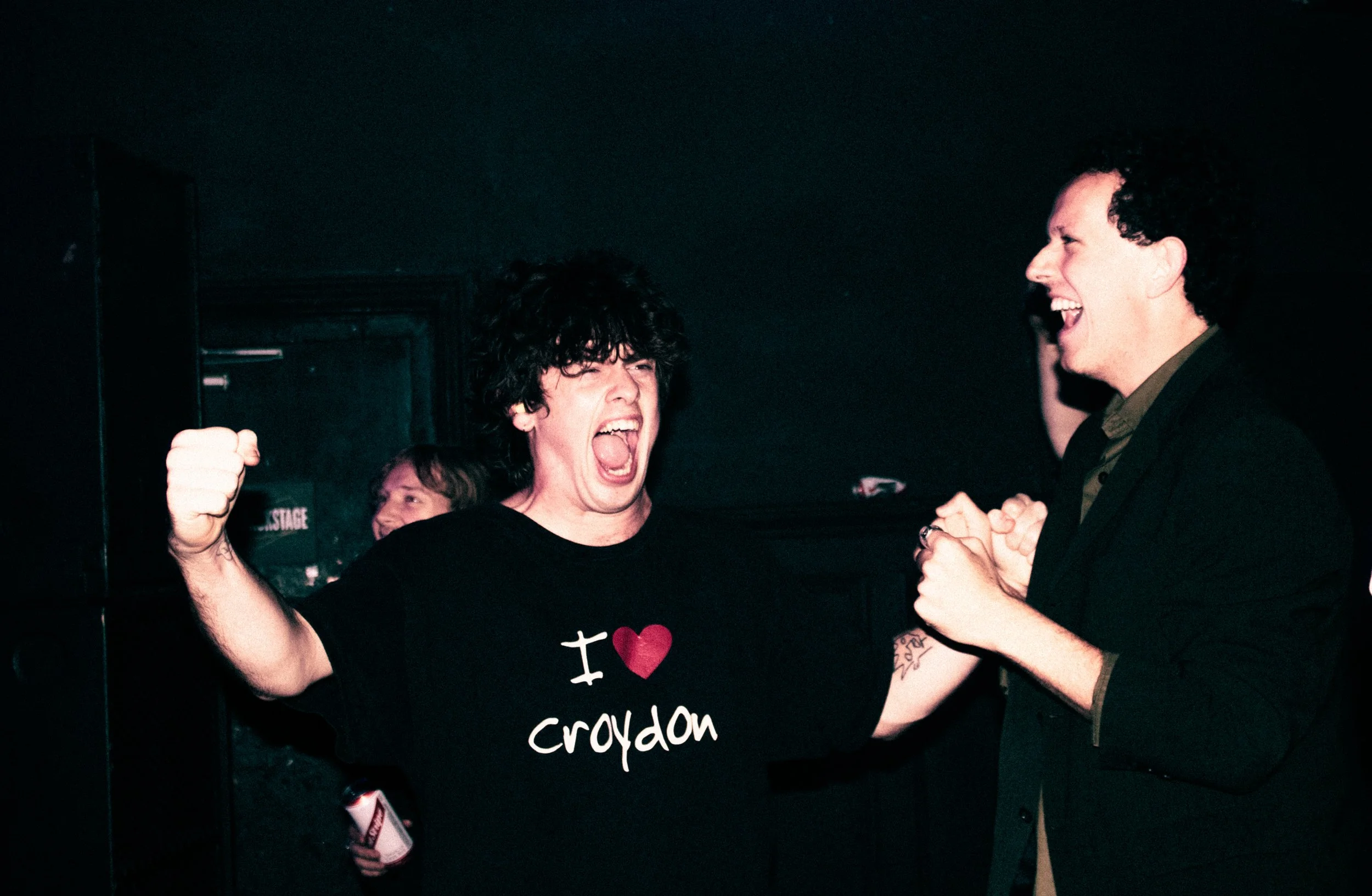 A woman with black curly hair wearing a black t-shirt that says 'I ❤️ croydon' is celebrating with her fist raised, enthusiastically speaking to a man in a suit who is smiling and clenching his hands in excitement.