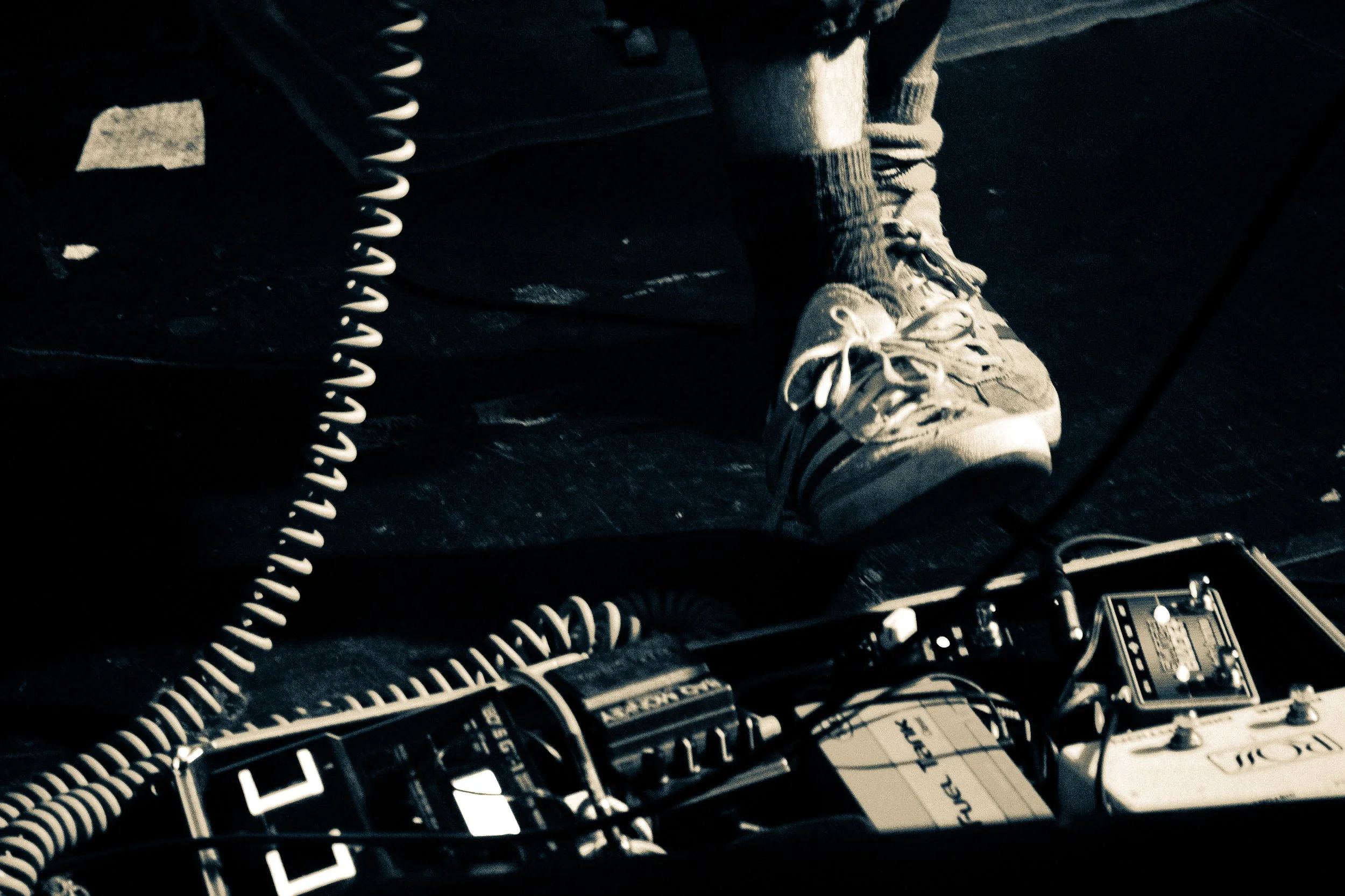 Close-up of a person's foot wearing a sneaker, resting on a pedalboard with effects pedals, in a dark setting, possibly during a musical performance.