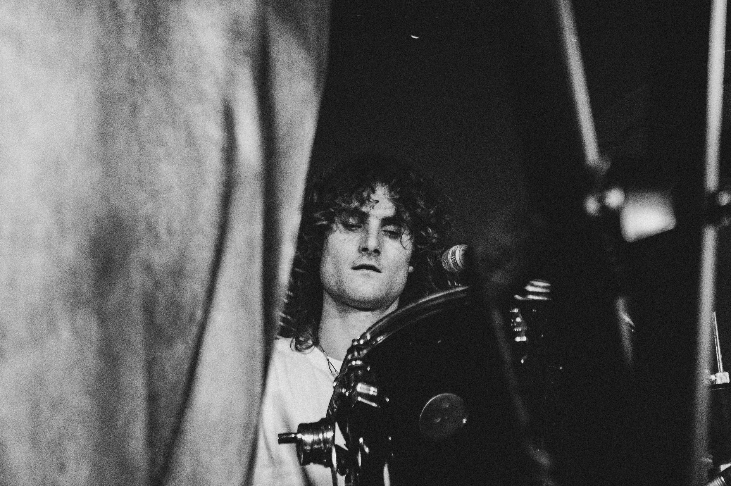 A black and white photo of a person with curly hair looking down at a camera or film equipment, partially obscured by a foreground object.