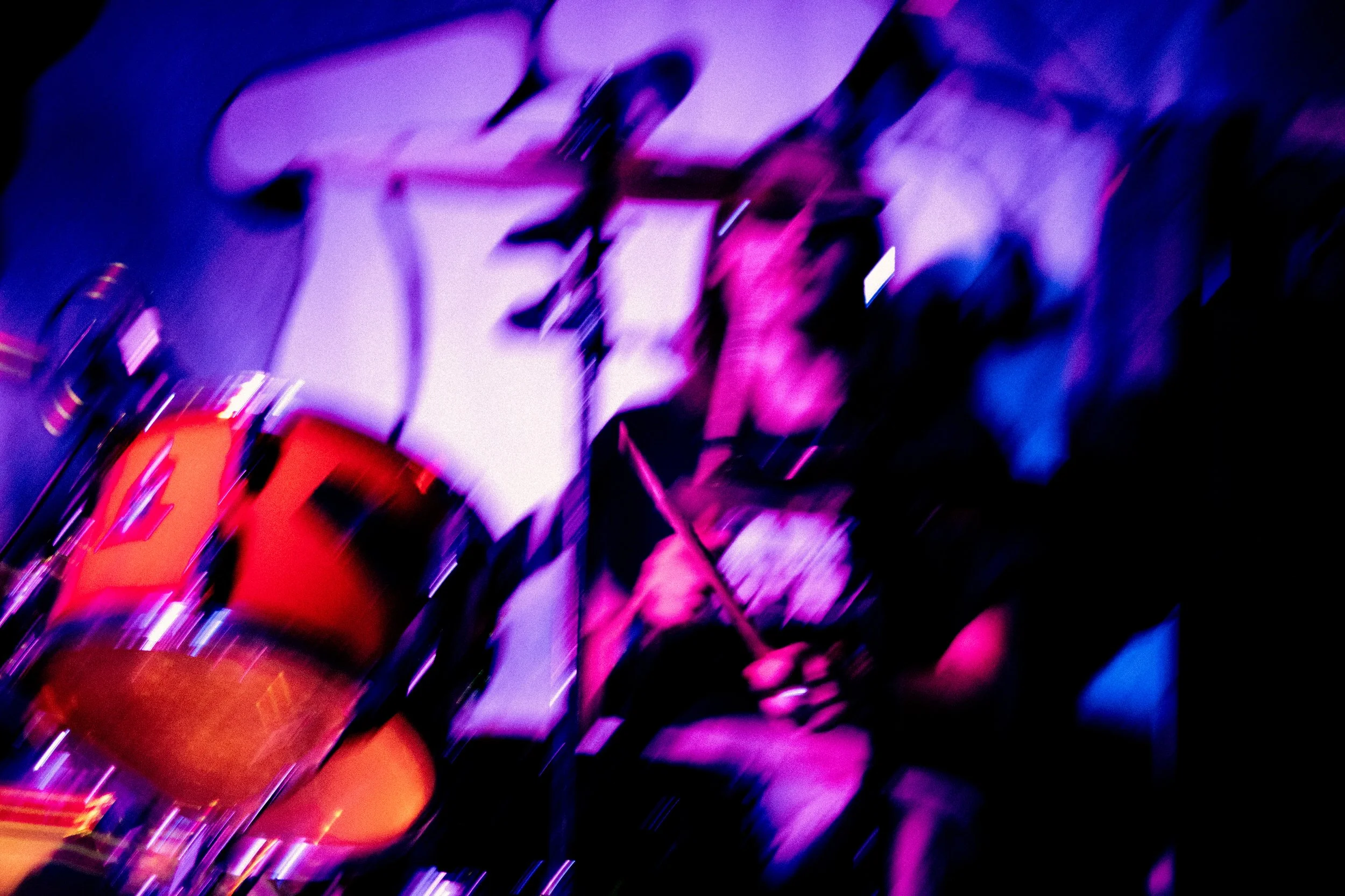 A blurry photo of a person playing drums, with purple and pink lighting, in a dark environment.