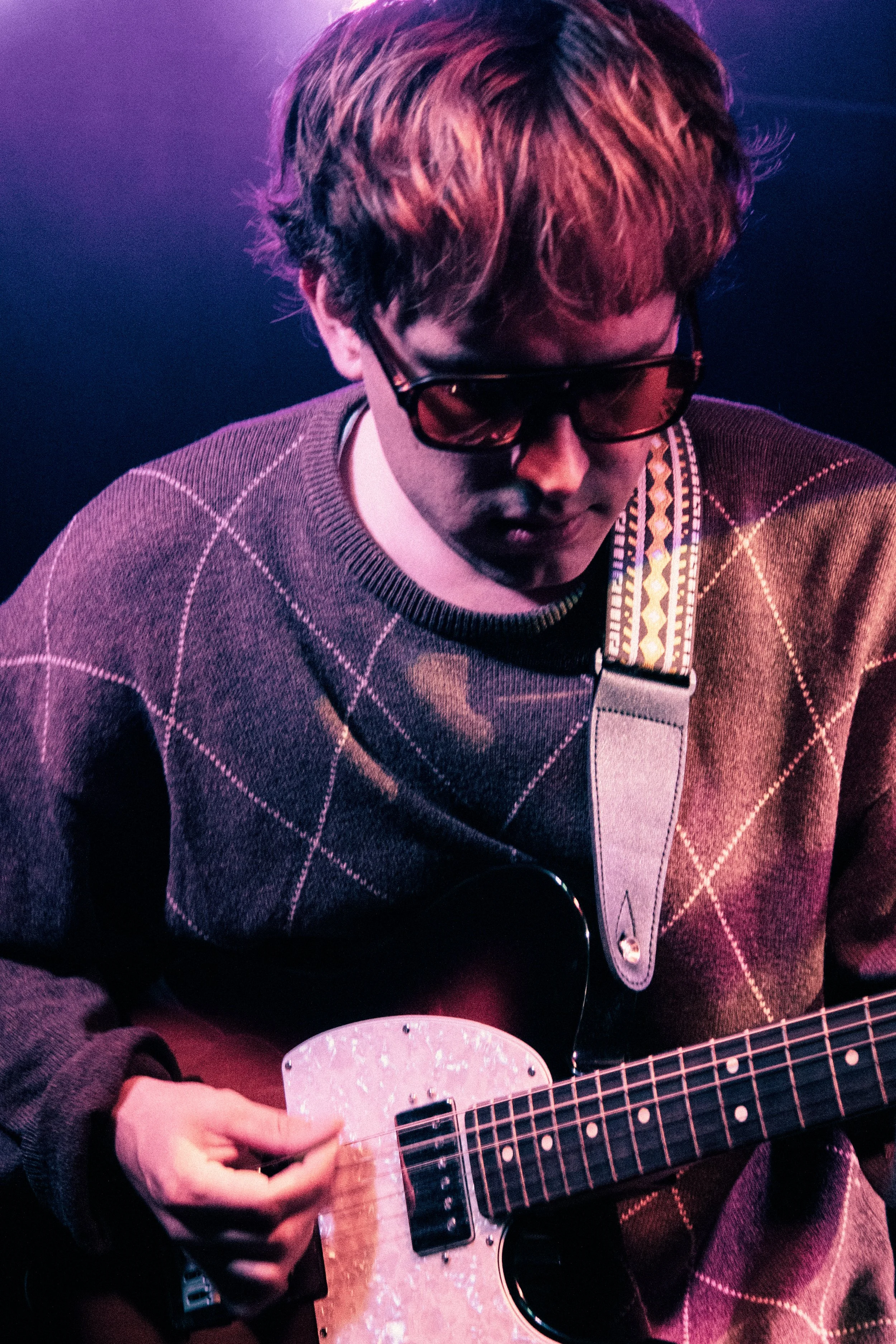 A young man wearing sunglasses and a checkered sweater is playing an electric guitar with a colorful strap in a dimly lit setting with purple and pink lighting.