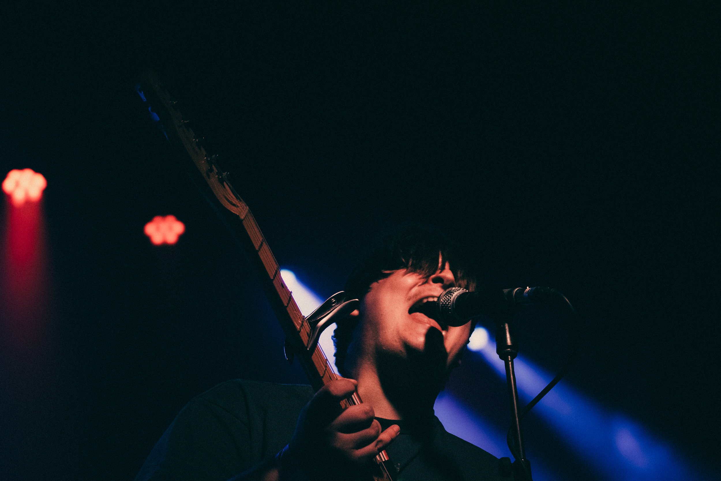 A person singing into a microphone while holding an electric guitar during a live performance in a dark setting with stage lights.