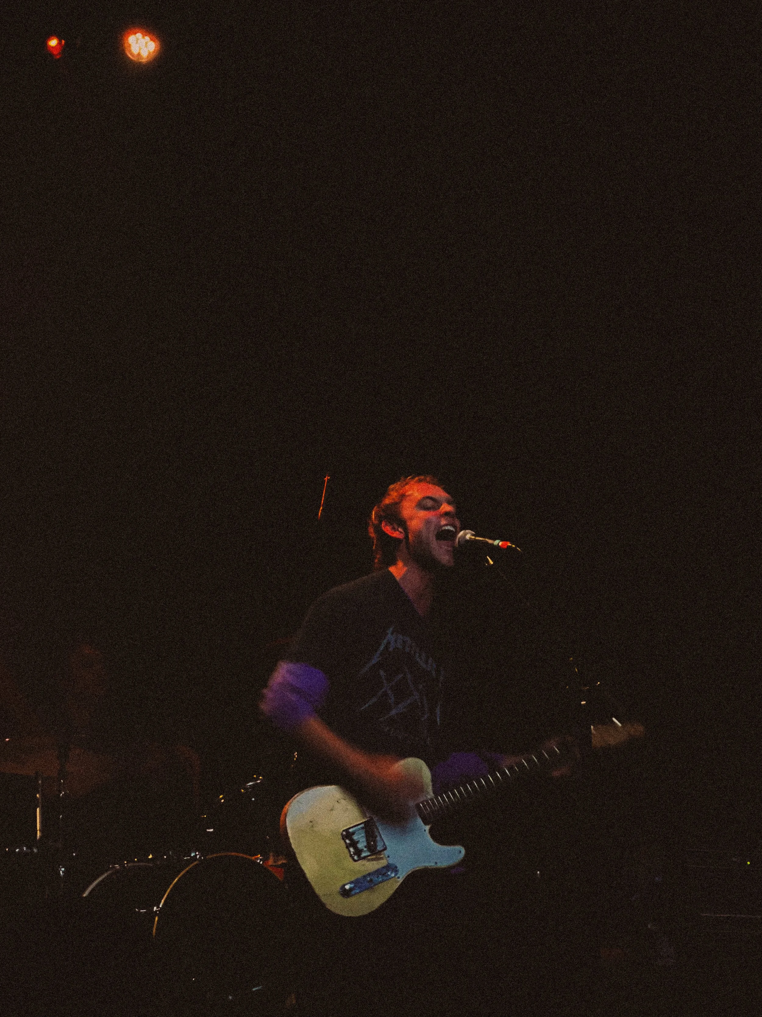 A male musician singing into a microphone and playing an electric guitar on stage during a live performance.
