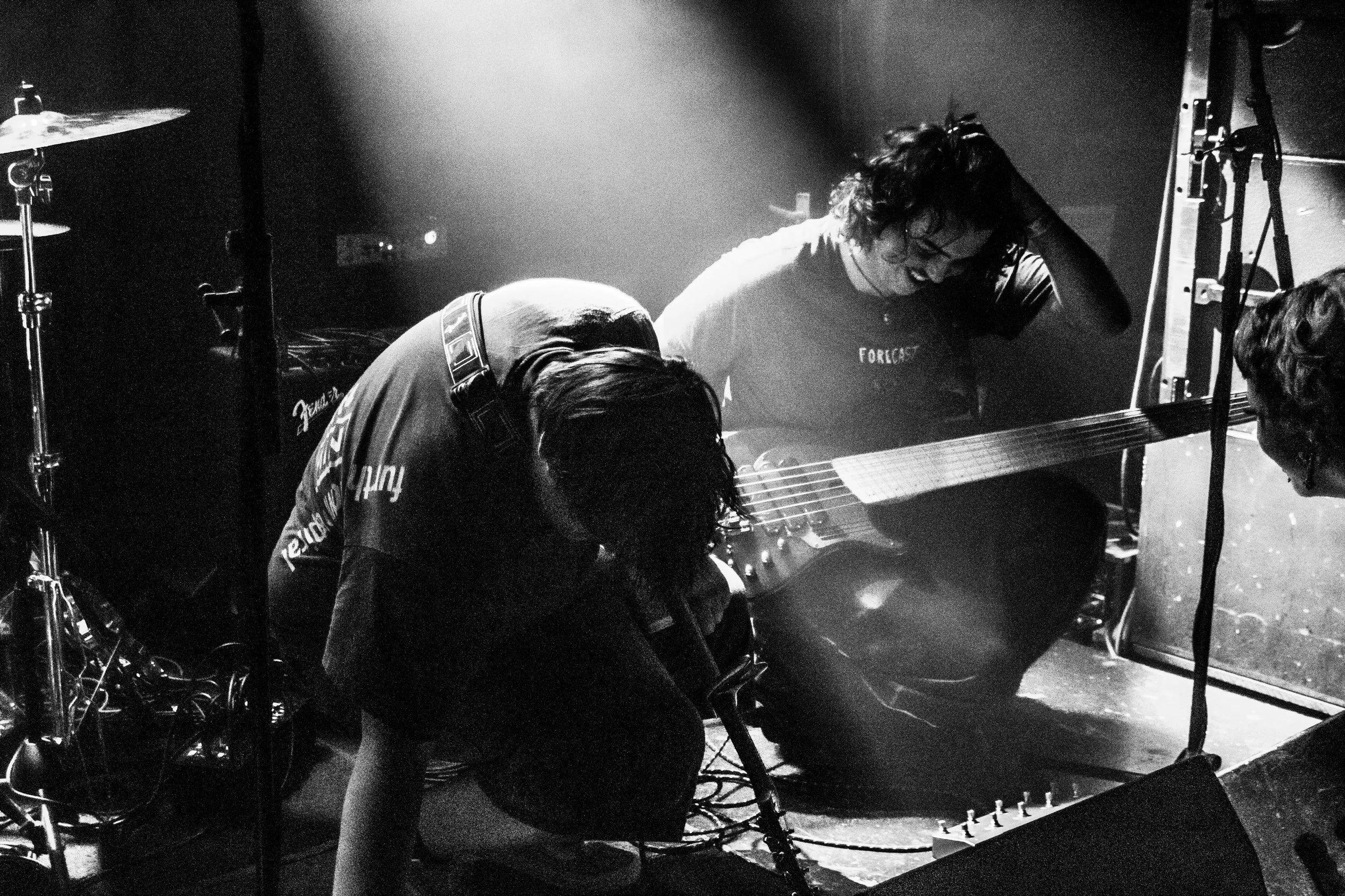 Two musicians on stage, one playing an electric guitar and the other sitting and singing into a microphone, with musical equipment around them in a dark concert setting.