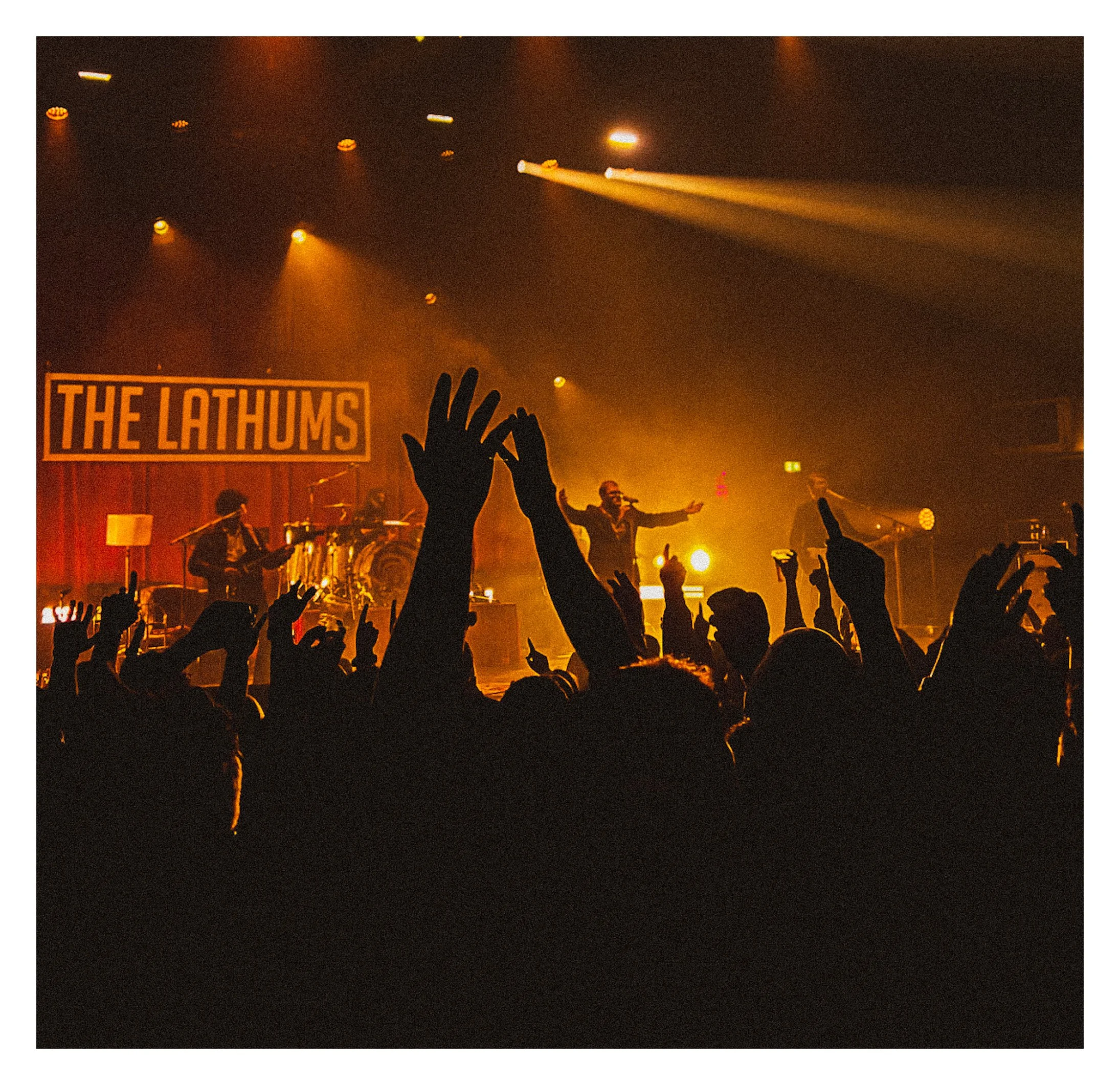 Concert scene with a band performing on stage and an audience raising their hands in front. The band has a sign that reads "The Lathums" in the background, and the stage is lit with yellow and orange lights. The Lathums at the 02 academy Brixton,Lond