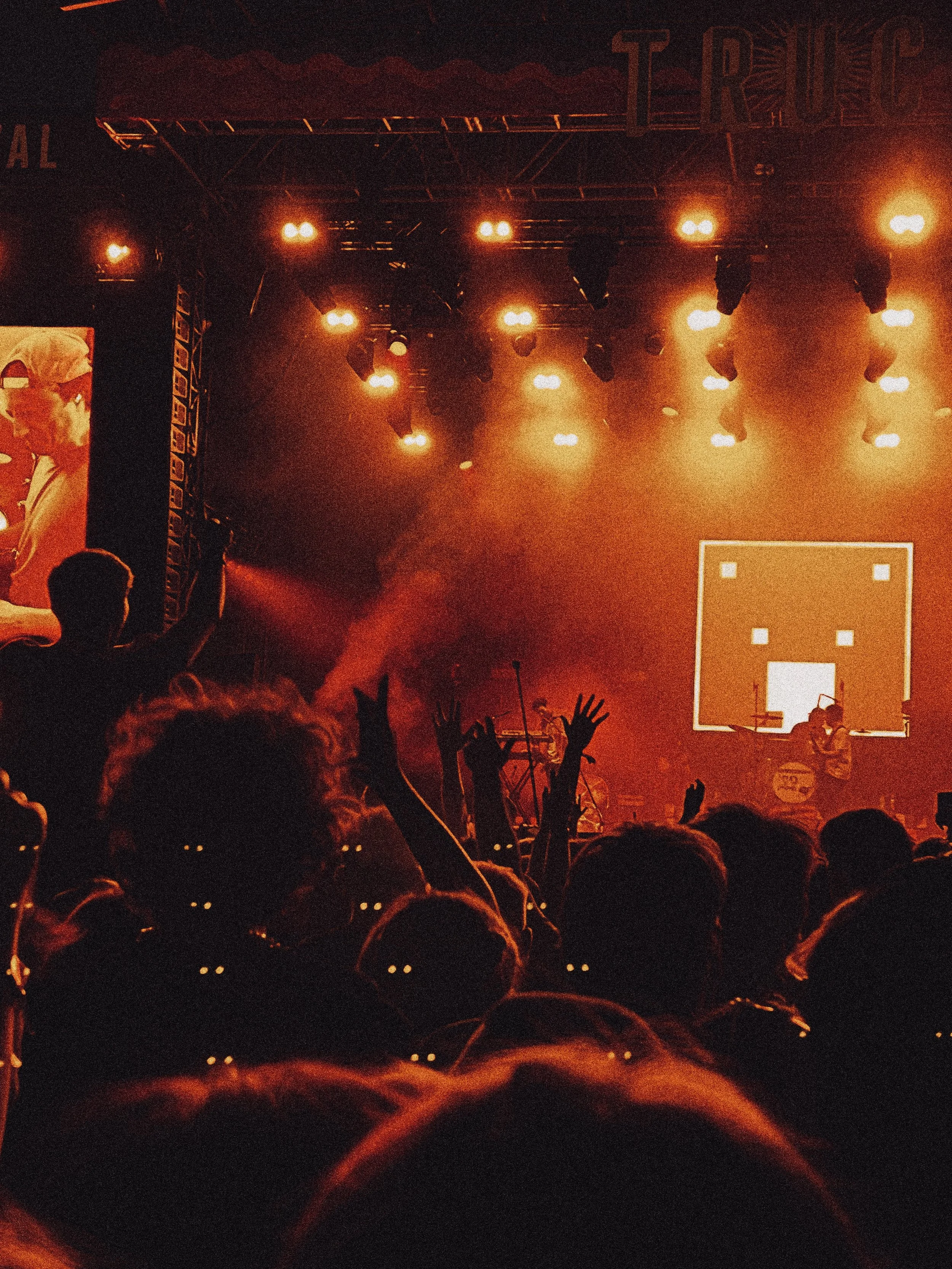 Crowd at concert with raised hands, stage with performers, bright orange lighting, and large screen on side