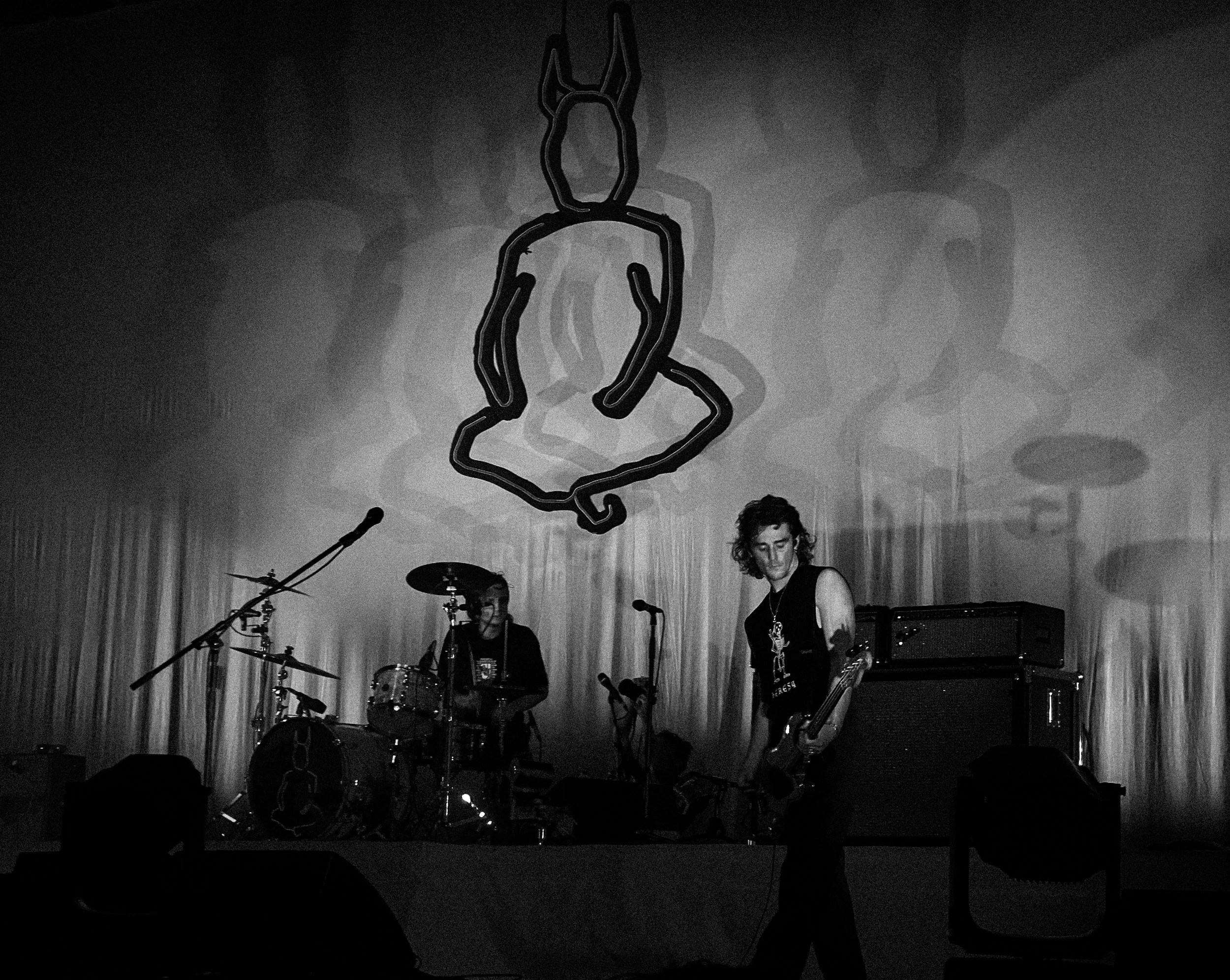 A black and white photo of a band performing on stage with a large neon outline of a rabbit behind them. The band has a drummer and a guitarist, and there are microphones and amplifiers on stage.