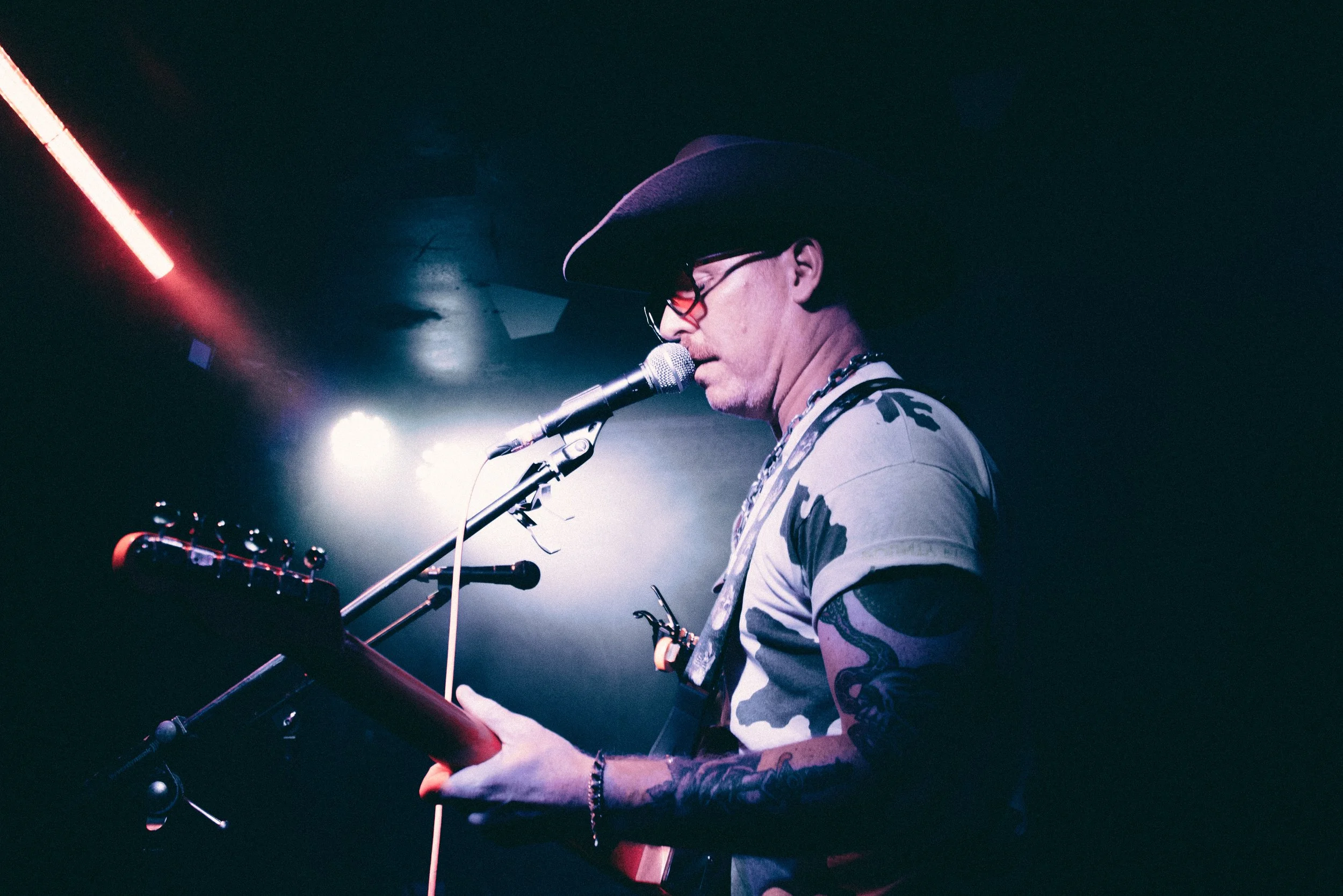 A man wearing a cowboy hat, glasses, and a camouflage t-shirt plays guitar and sings into a microphone on a dimly lit stage with bright lights behind him.