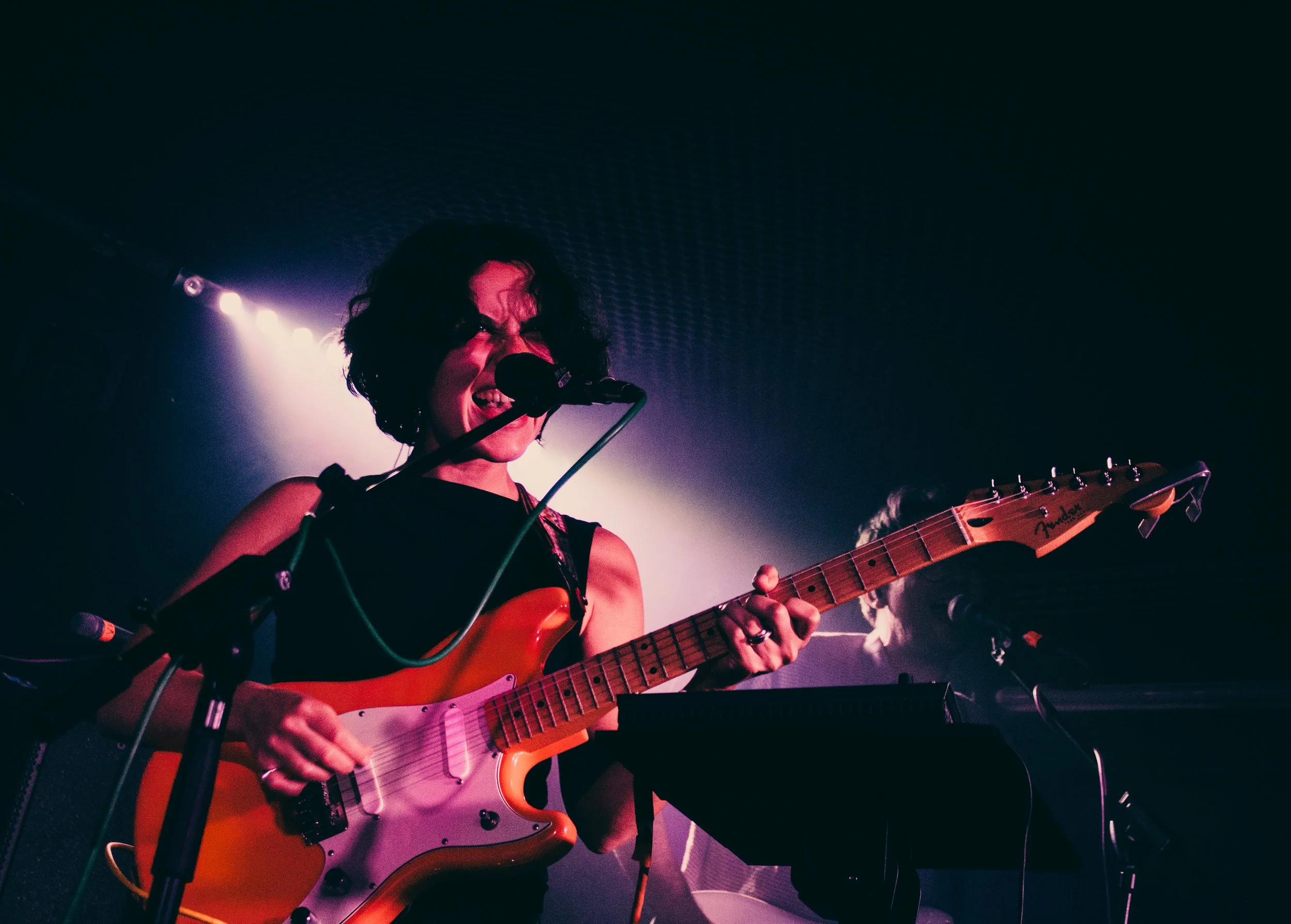 Woman with short dark hair singing into a microphone and playing an orange Fender electric guitar in a music performance with purple lighting. Lazy Day.