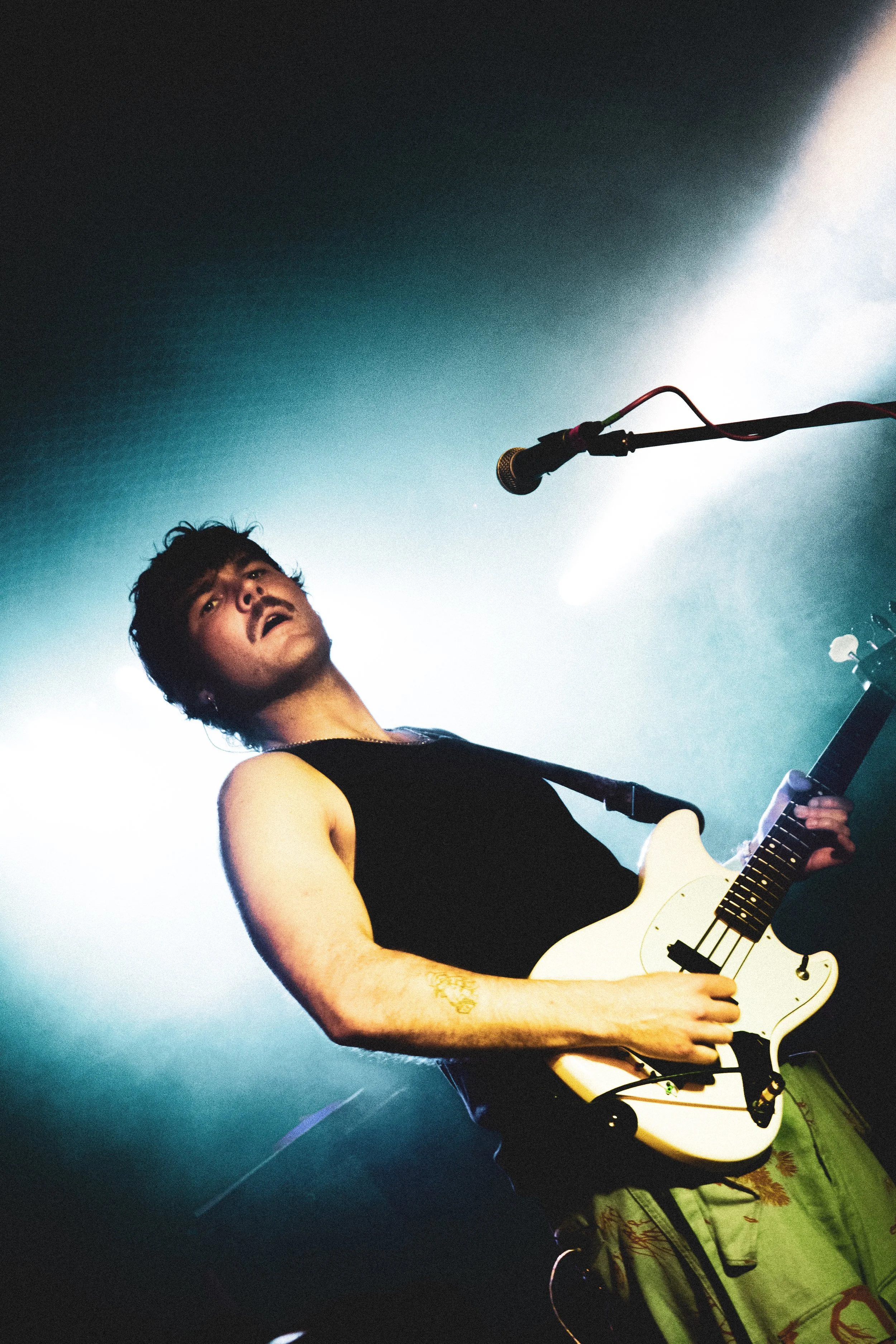 A male guitarist performing on stage with a microphone overhead, wearing a black tank top and green cargo pants, under concert lighting.