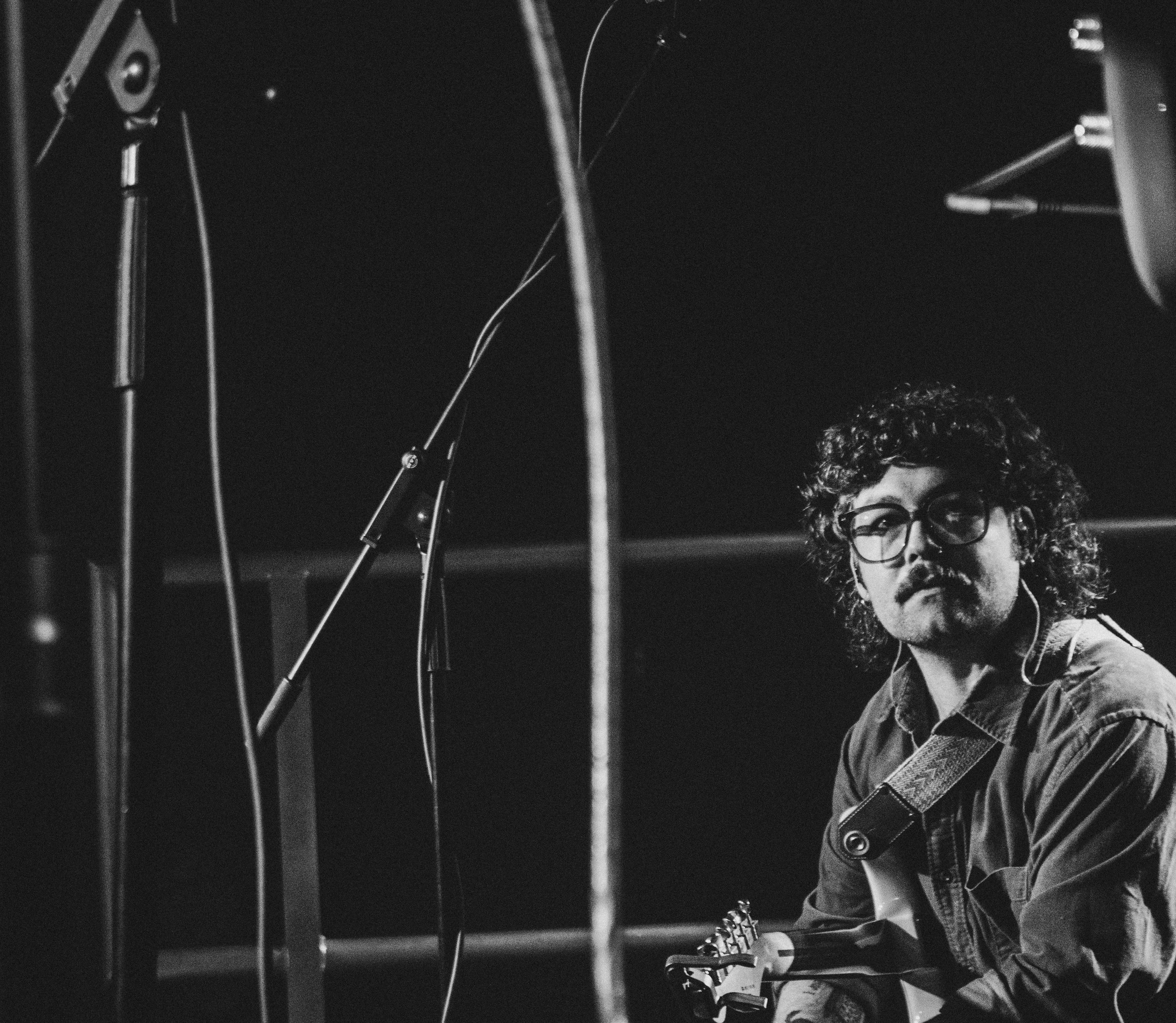 A man with curly hair, glasses, and a mustache, holding a guitar, seated in a recording studio with microphone stands and recording equipment around him, in black and white.