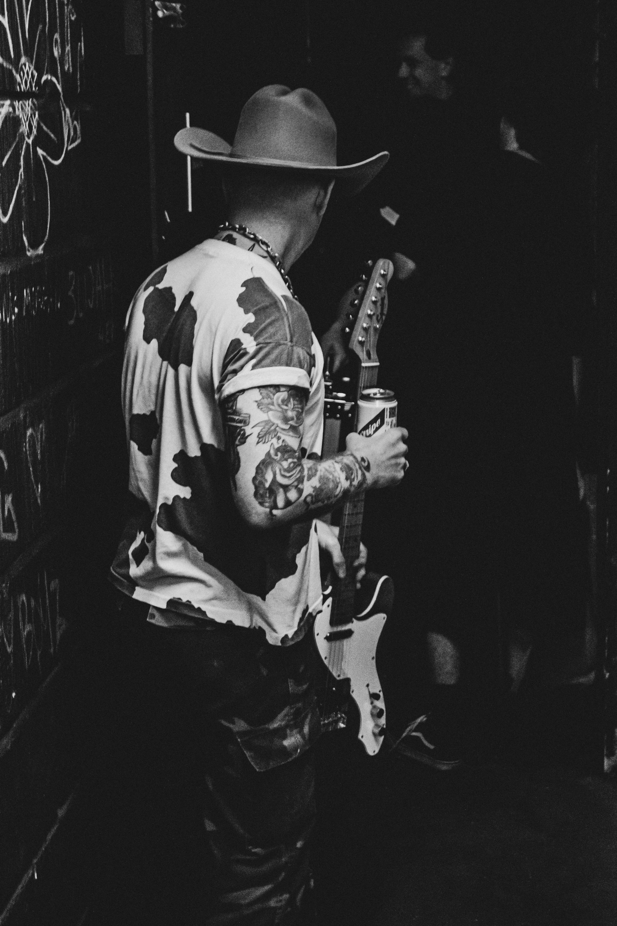 A man with tattoos on his arm wearing a cowboy hat and a cow print shirt holding a guitar and a can of beer, standing in a dimly lit room.