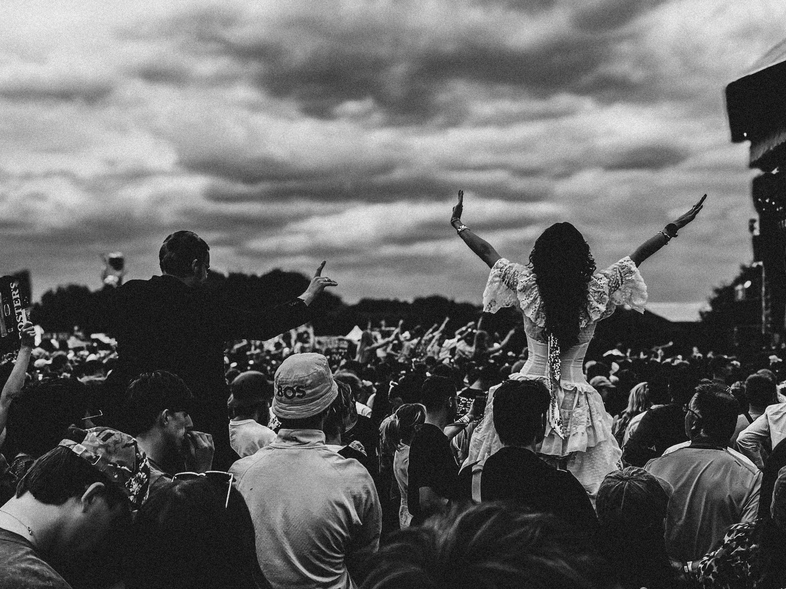 A large crowd at an outdoor event under dark, cloudy skies. In the center, a woman in a white dress with her arms raised and a man next to her with one arm raised; the crowd is diverse, and people are watching or taking photos.