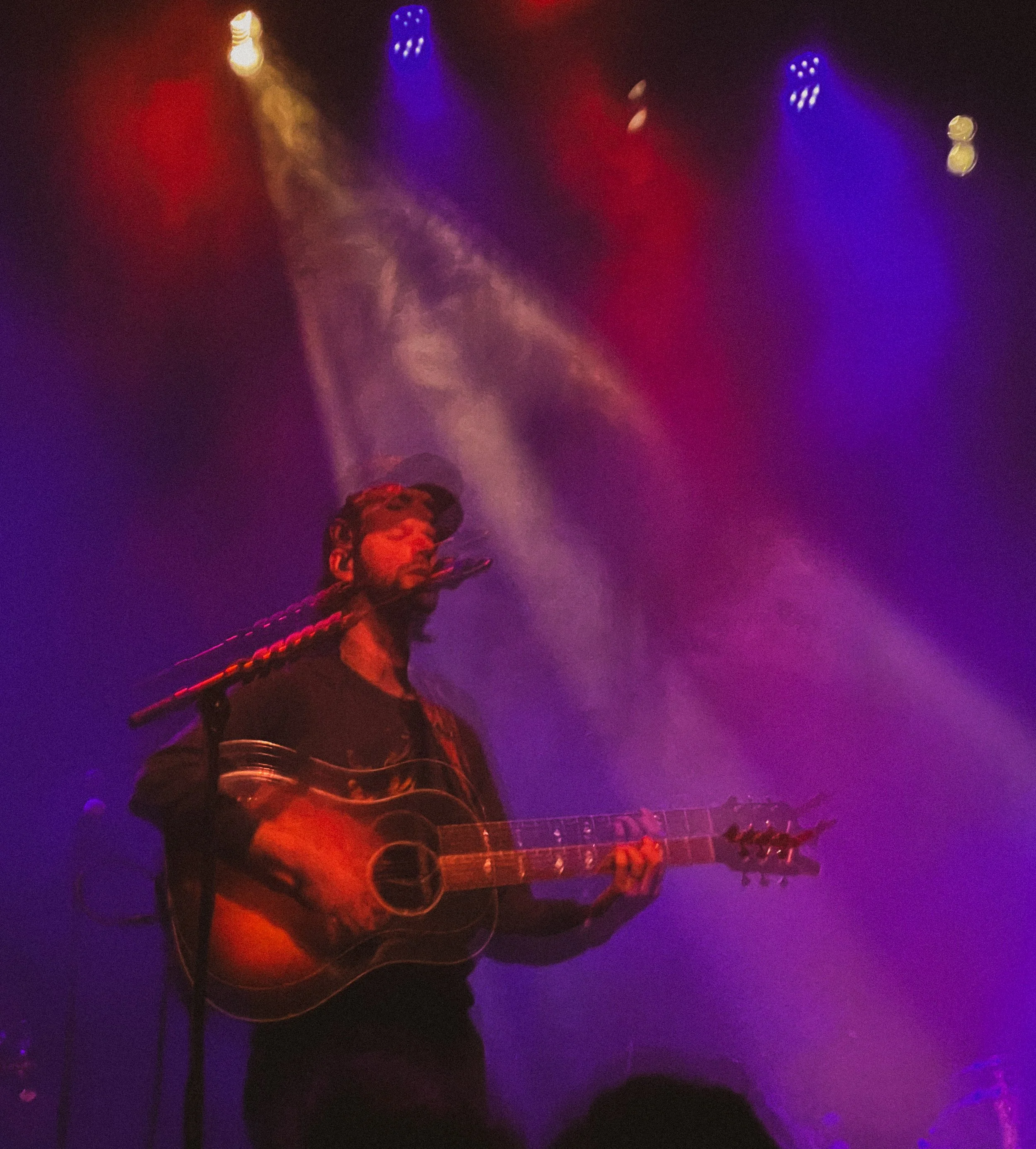 A musician playing an acoustic guitar on stage at a concert, with colorful stage lighting including red, yellow, and purple hues.
