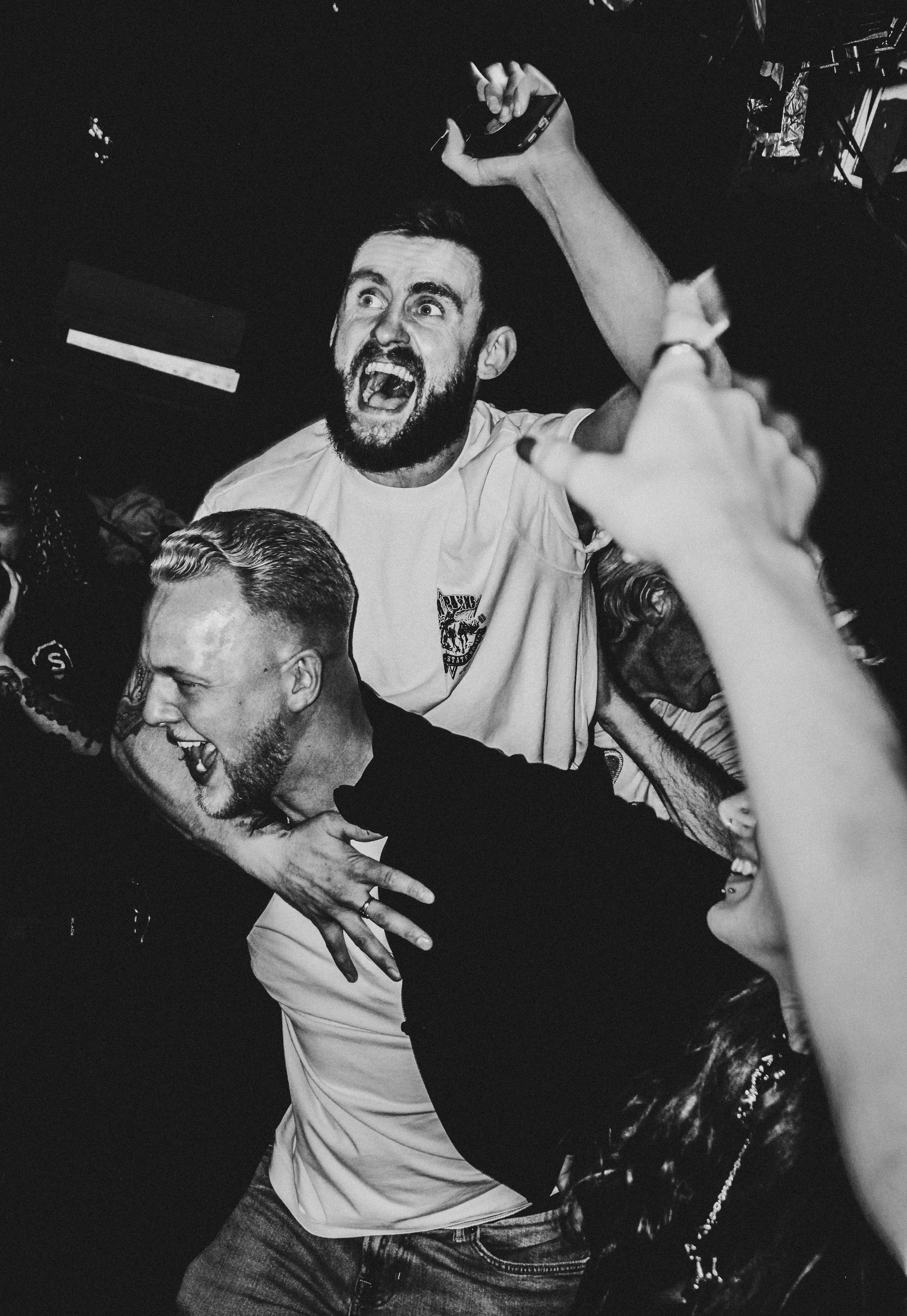 A group of people celebrating and having fun at a party, with one man enthusiastically shouting and raising his arm, while another man is being playfully restrained, and a woman is smiling in the foreground. TEZ band headline in London, crowd. Energe