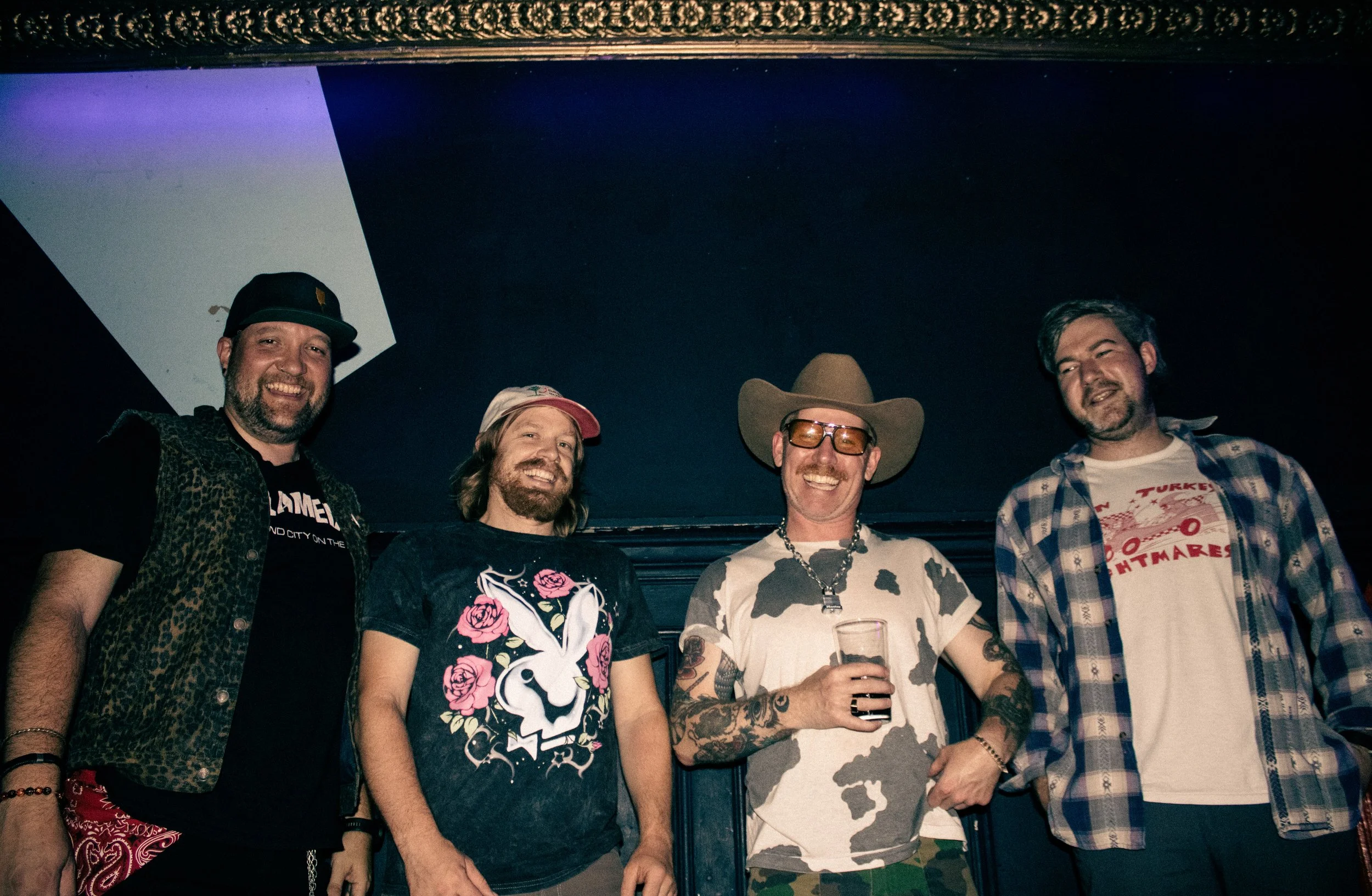 Four men standing together, smiling at the camera, in a dark indoor setting with a blue and purple background. The man on the far left is wearing a black cap and a black t-shirt with a leopard print vest. The second man from the left has long hair, a
