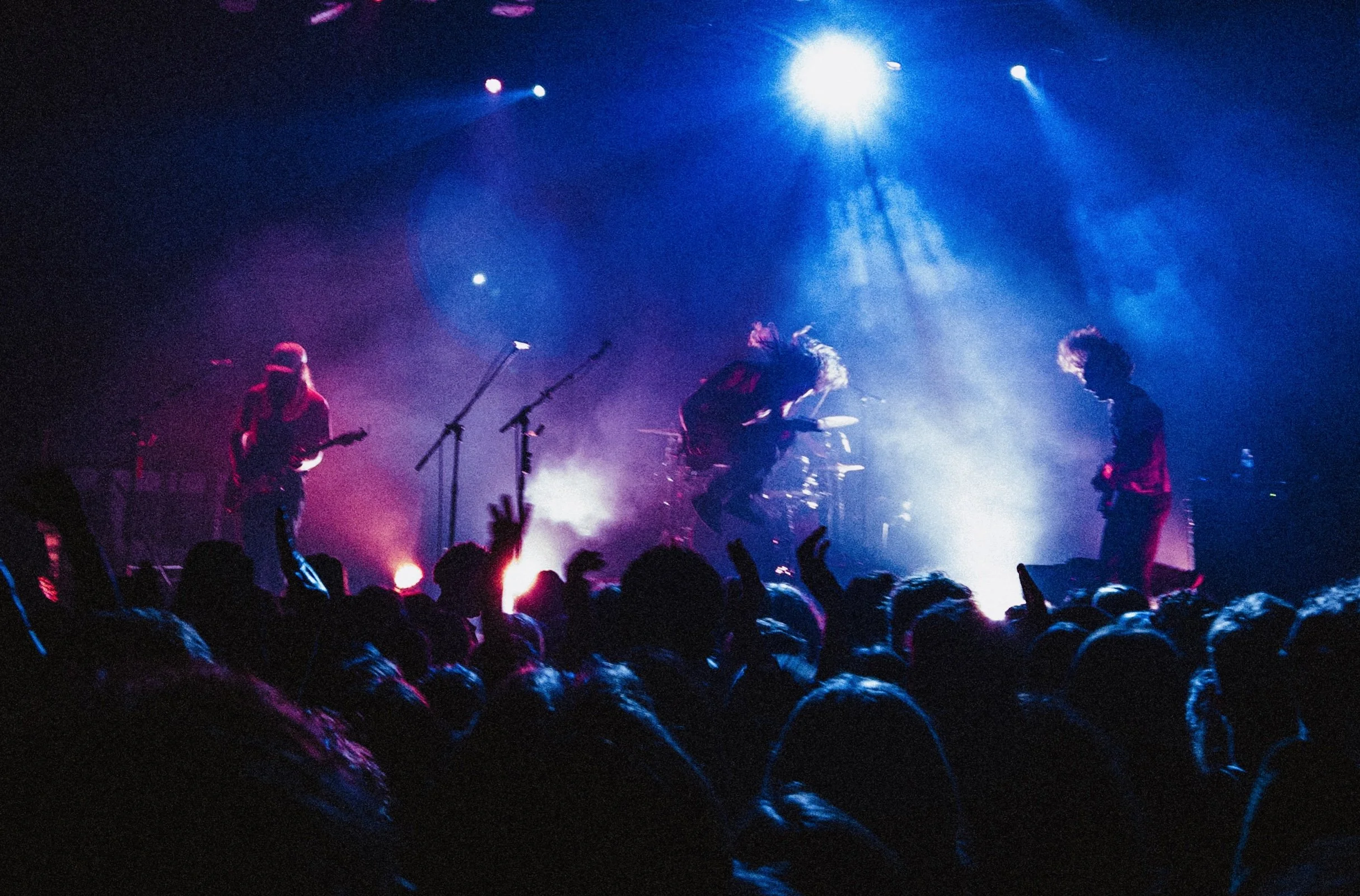 A band performs on stage with bright lights and a fog machine, and a crowd watches.