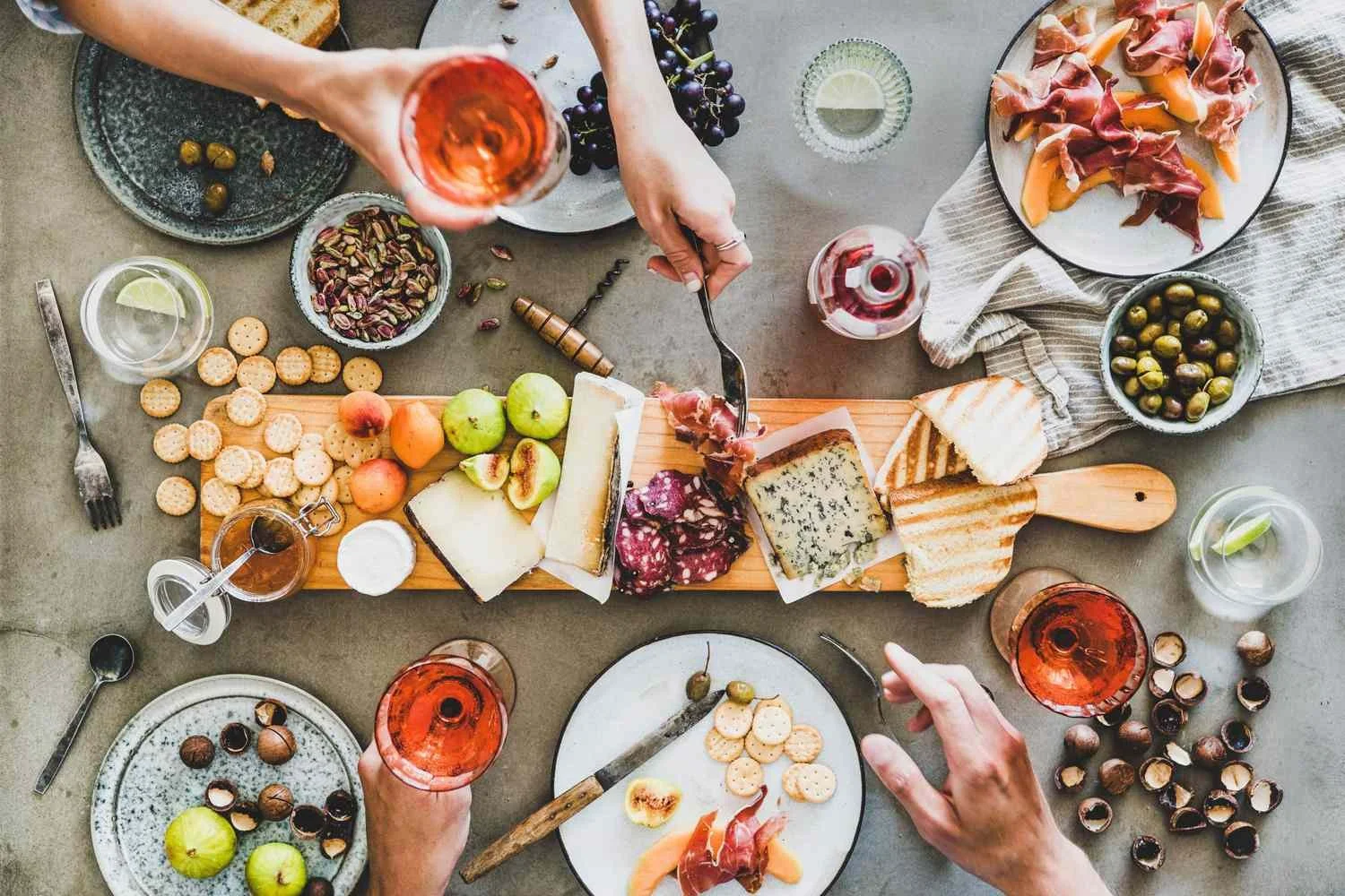 Top-down view of a charcuterie board with cheese, fruits, crackers, and meats, surrounded by glasses of rosé wine, on a gray table with additional dishes and utensils.