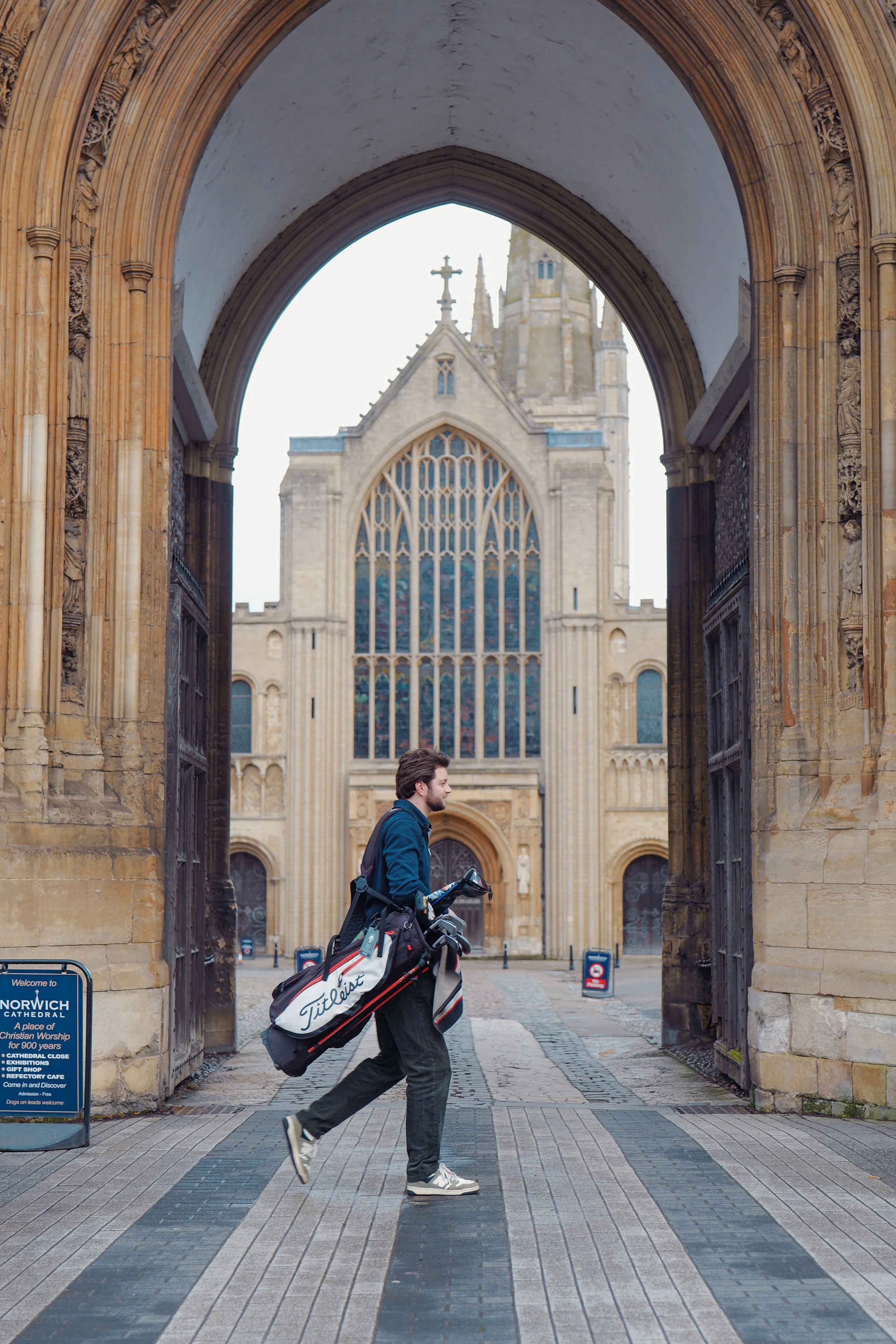 A man with a golf bag walking through the opening of a historic church, Normandy Cathedral, with gothic architecture and stained glass windows, in the background.