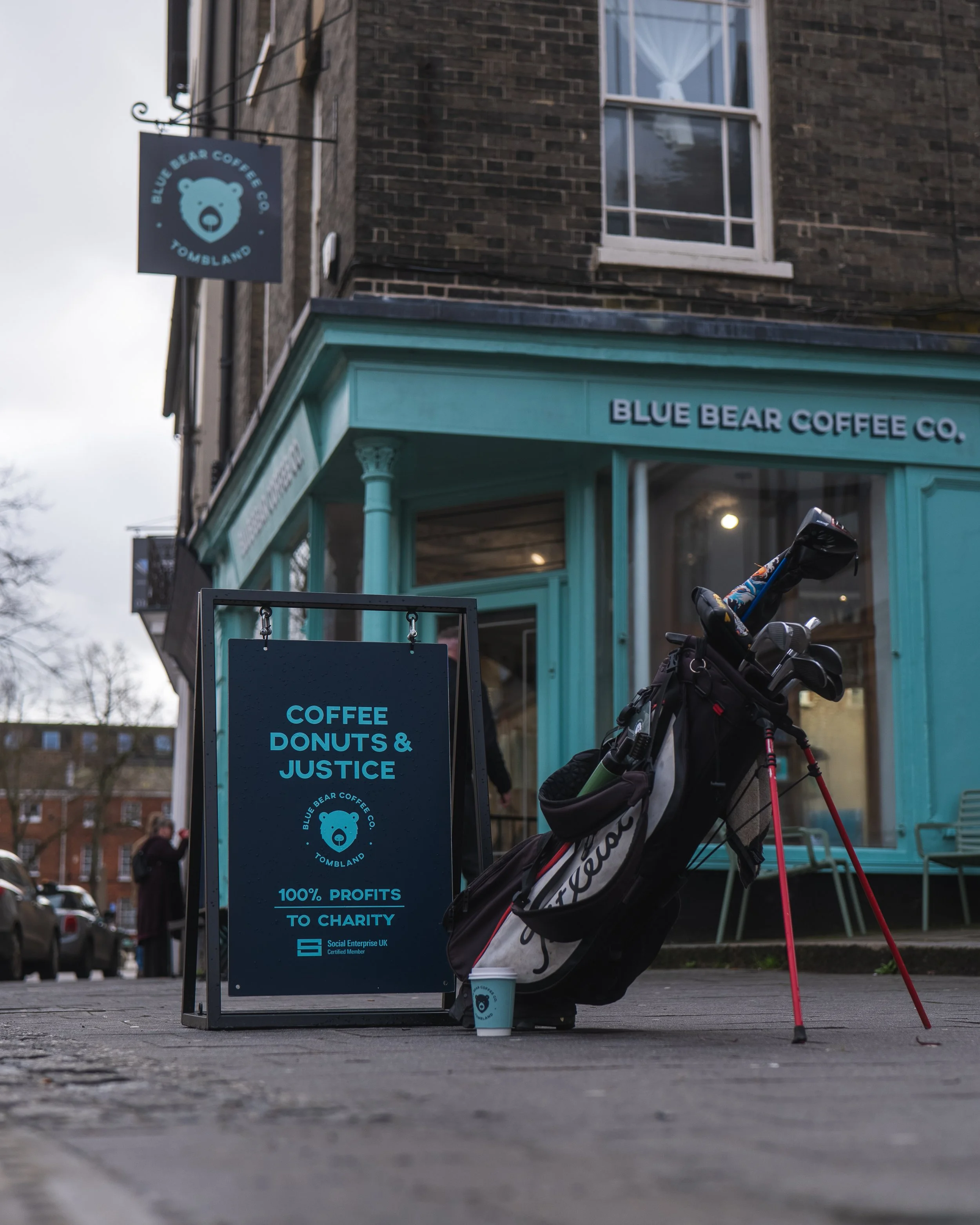 A street view of Blue Bear Coffee Co. with a sidewalk sign that reads 'Coffee, Donuts & Justice. 100% Profits to Charity.' A golf bag with clubs, a small coffee cup, and a person's feet are visible in front of the shop's teal-painted corner with larg