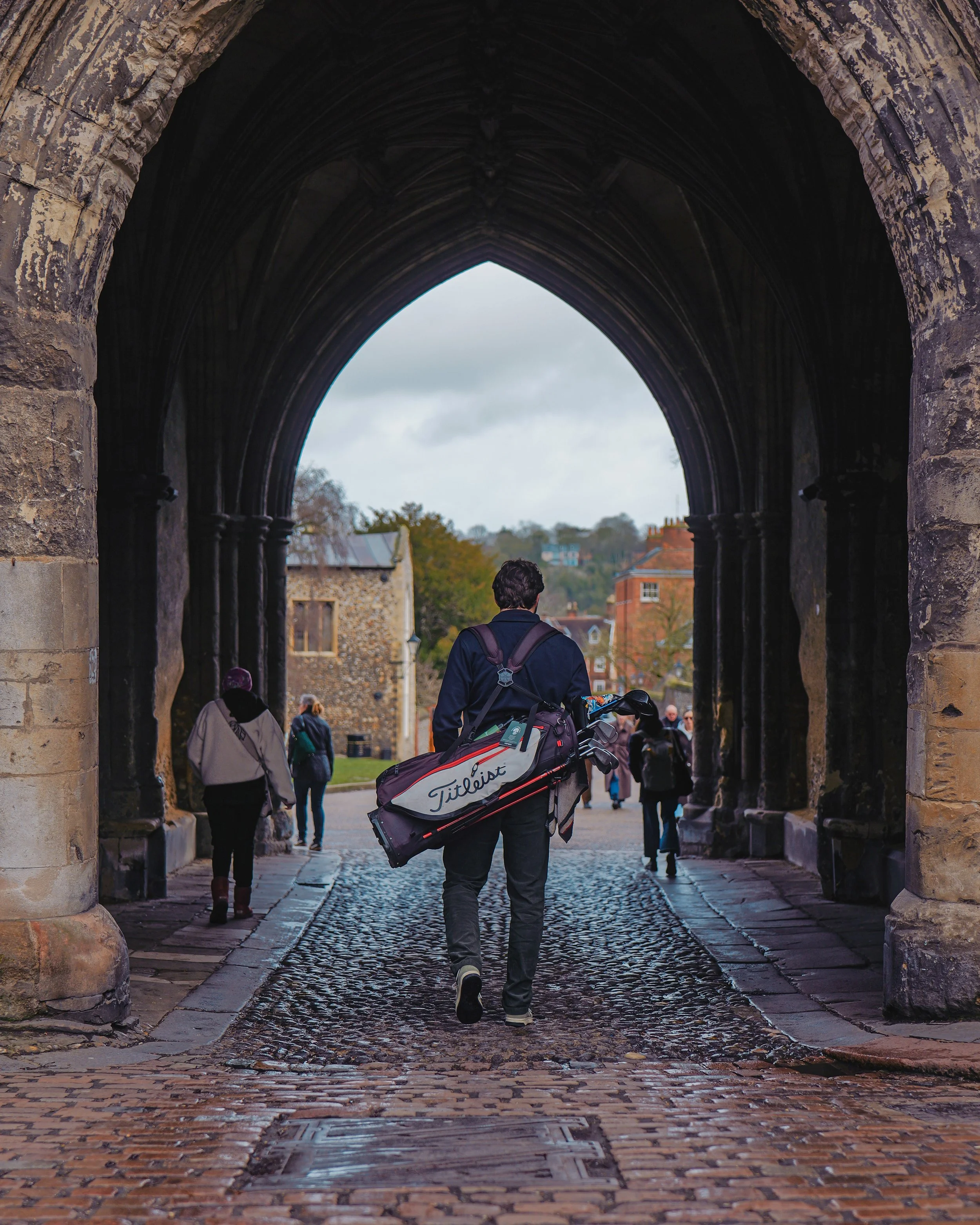 A man walking through a historic stone archway carrying a golf bag, with other pedestrians in the background and a cloudy sky.