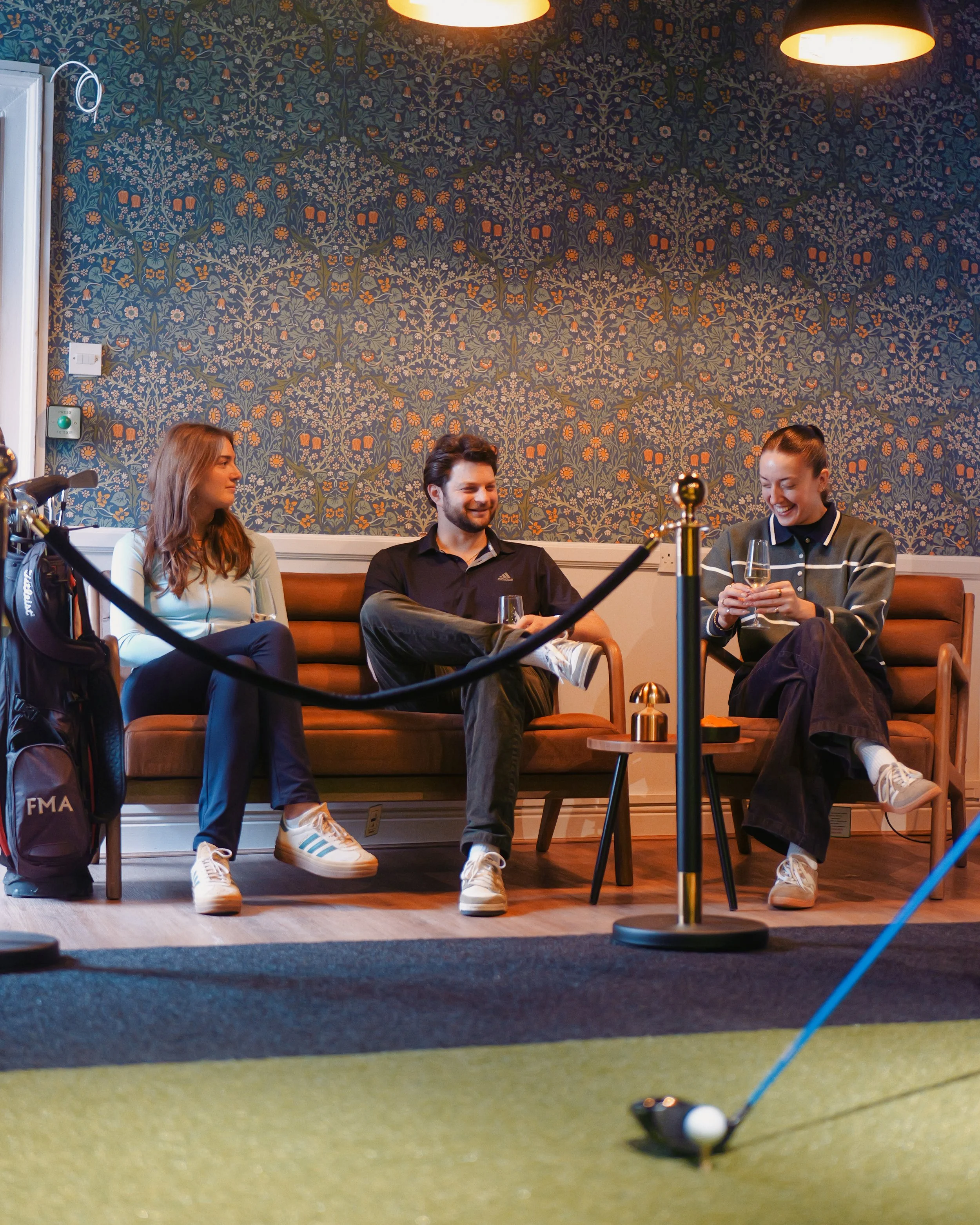 Three friends sitting on a sofa in a cozy, well-lit lounge area, laughing and talking, with a golf club and ball on a small indoor putting green in front of them.