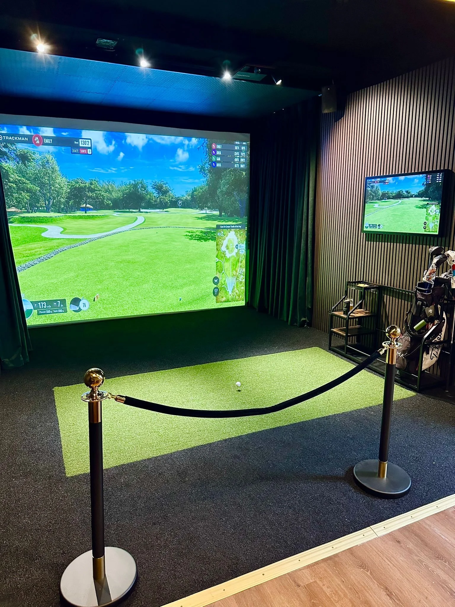 Indoor golf simulation room with a screen showing a virtual golf course, a golf ball on a small mat, and golf clubs on a stand.