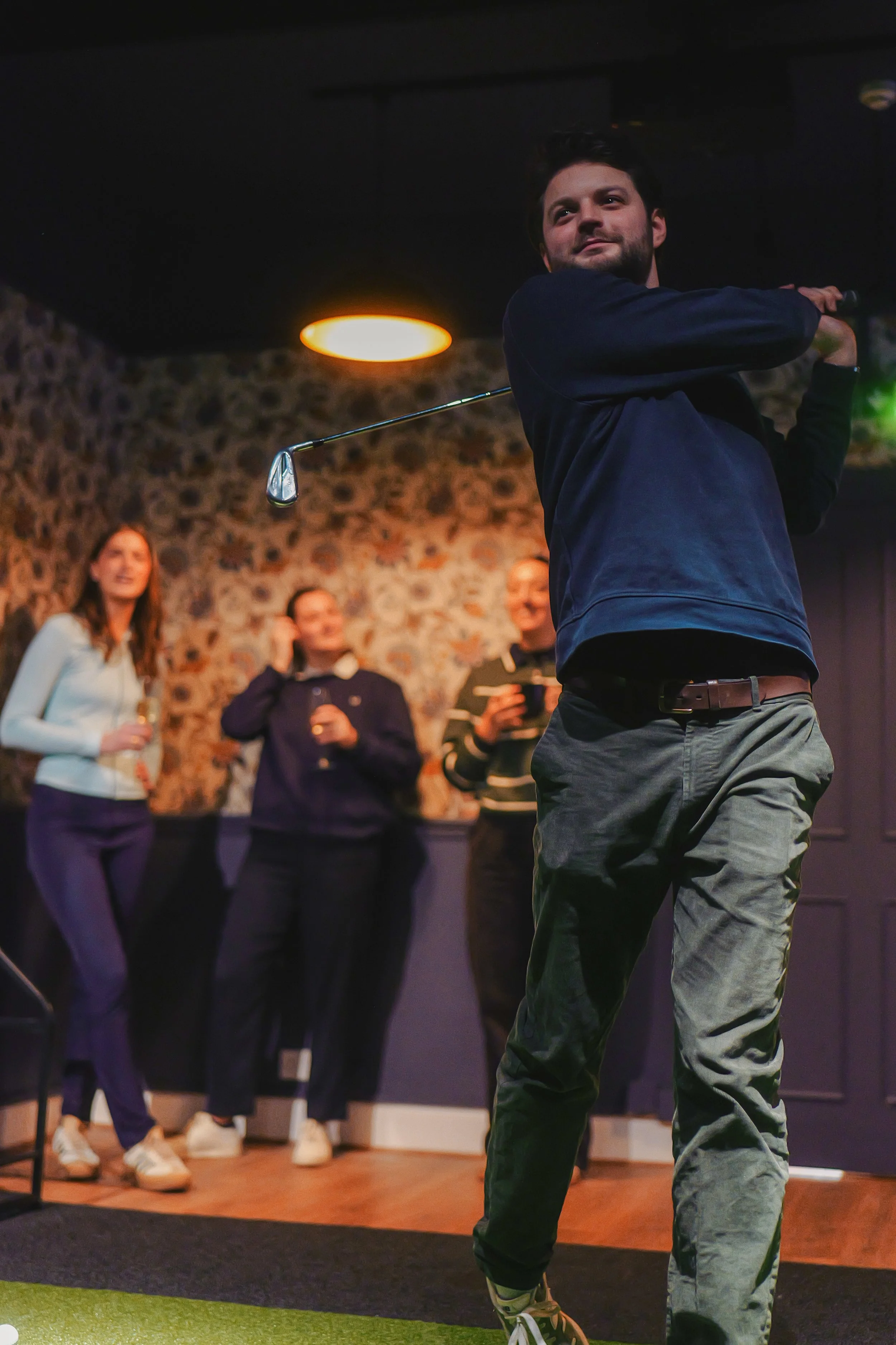 A man is swinging a golf club indoors at a gathering, with three women watching and smiling in the background.