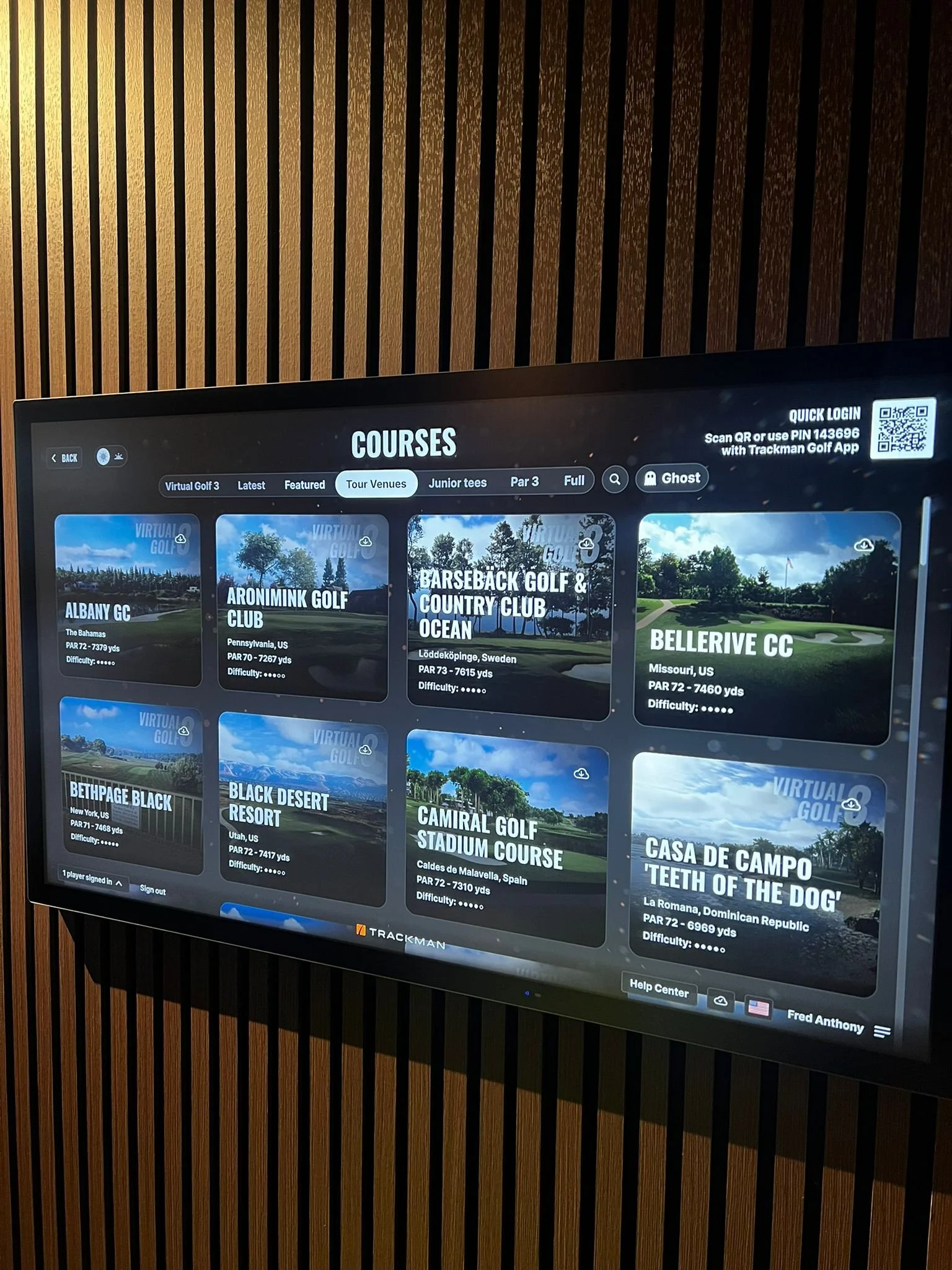 Digital screen displaying list of golf courses at a golf facility, including names, locations, and distances, with categories such as Virtual Golf, Latest, Featured, Tour Venues, Junior Tees, Par 3, and Full, mounted on a wall with vertical wooden paneling.