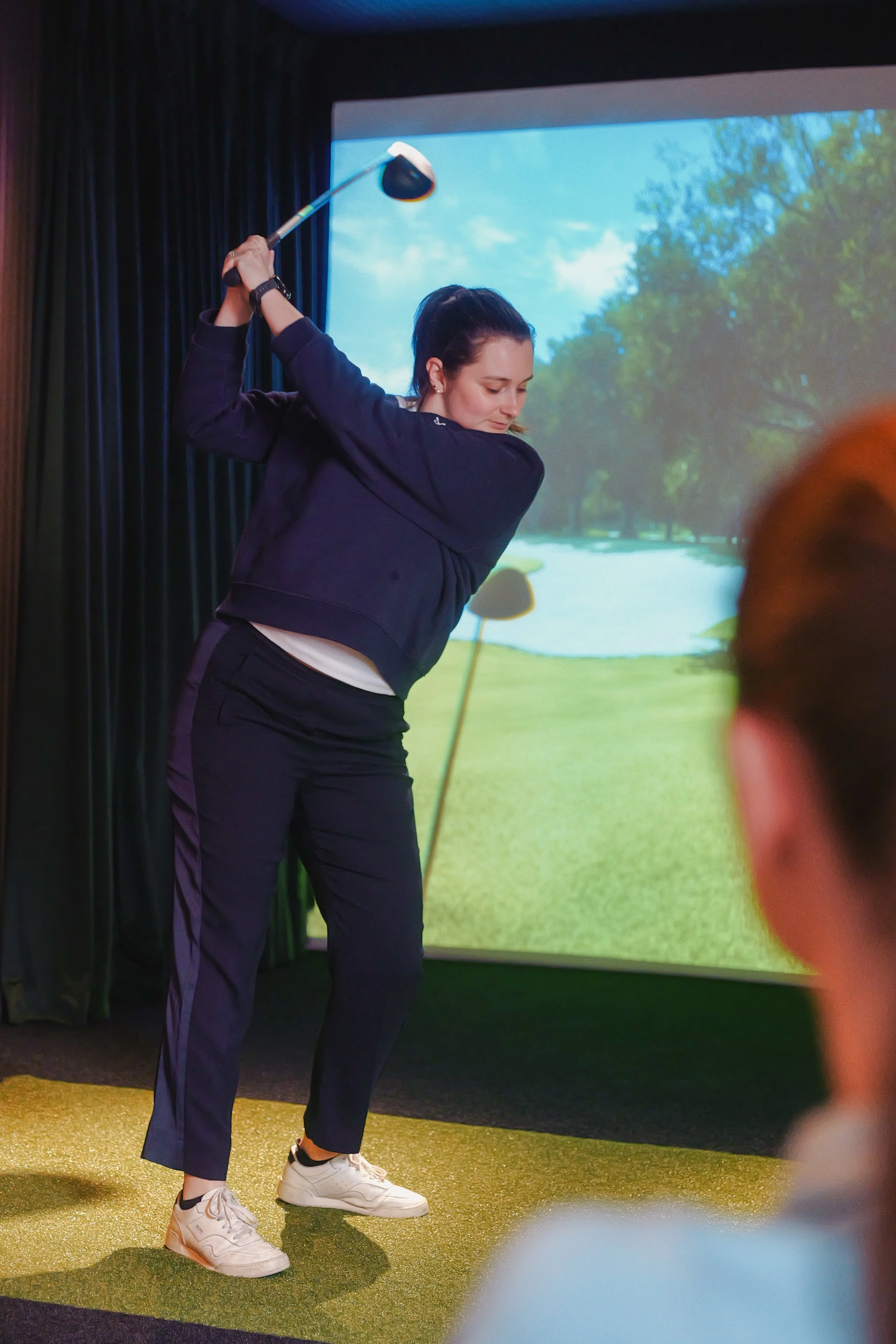 A woman practicing golf indoors, swinging a golf club in front of a golf course background projection.