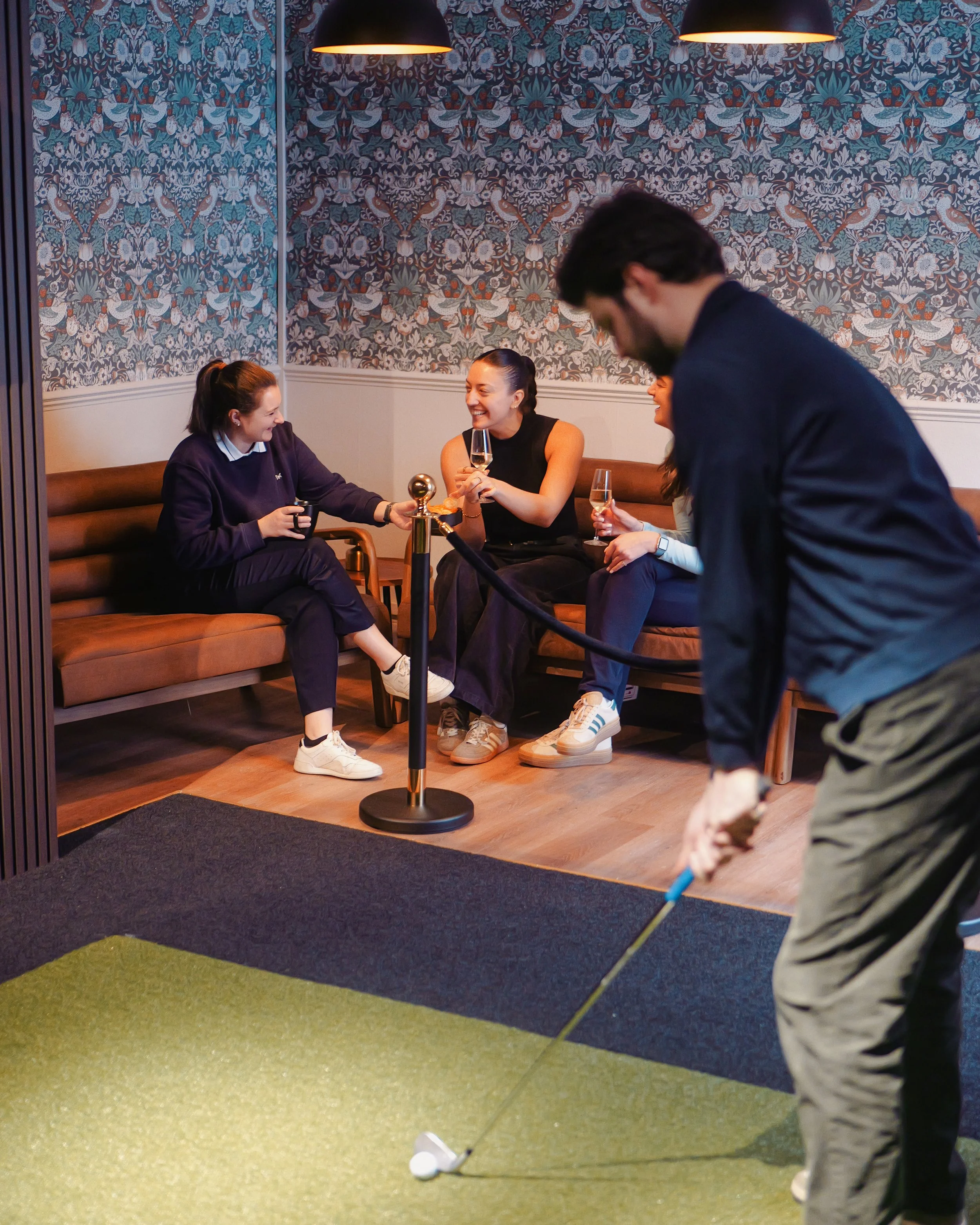 Four women sitting on a sofa in a lounge area, two holding glasses of champagne and chatting, while a man plays golf on an indoor putting green in front of them.