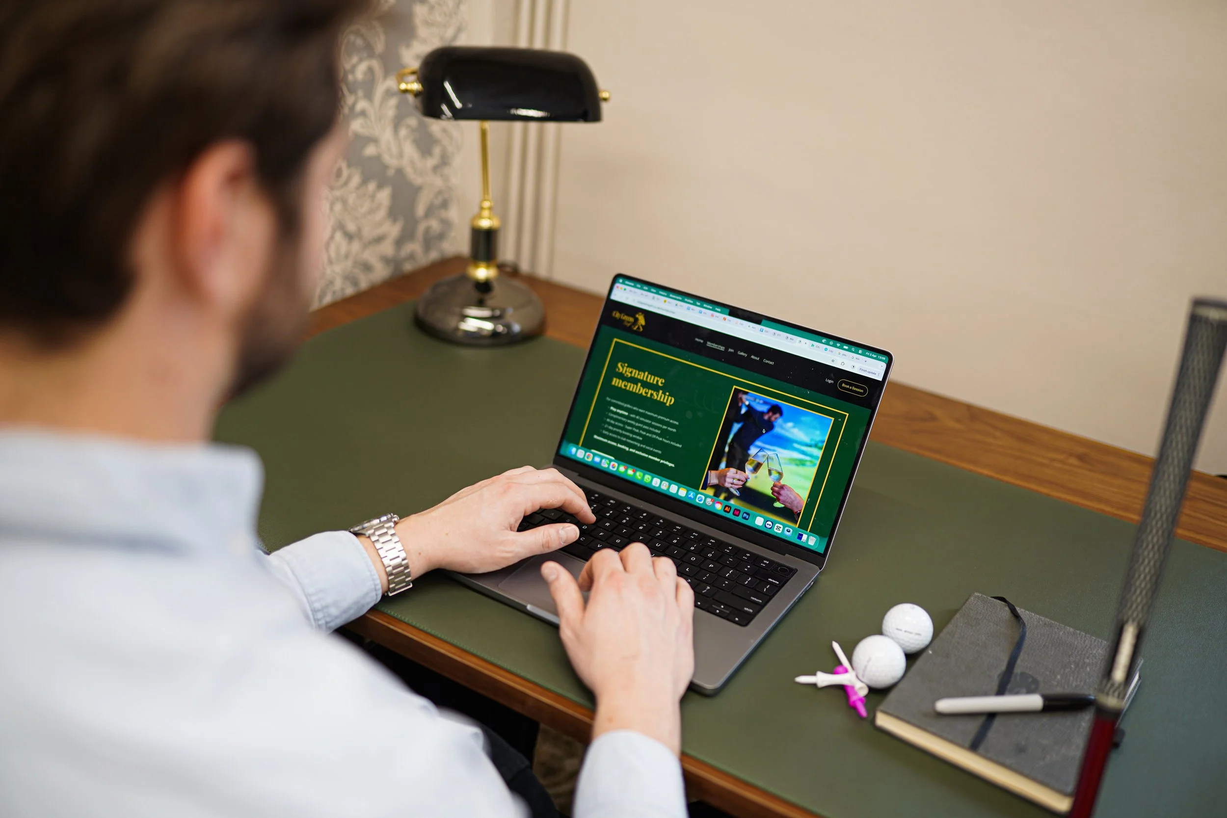 A person using a laptop displaying a website about golf membership, sitting at a desk with a green surface, a black desk lamp, golf balls, and golf tees.