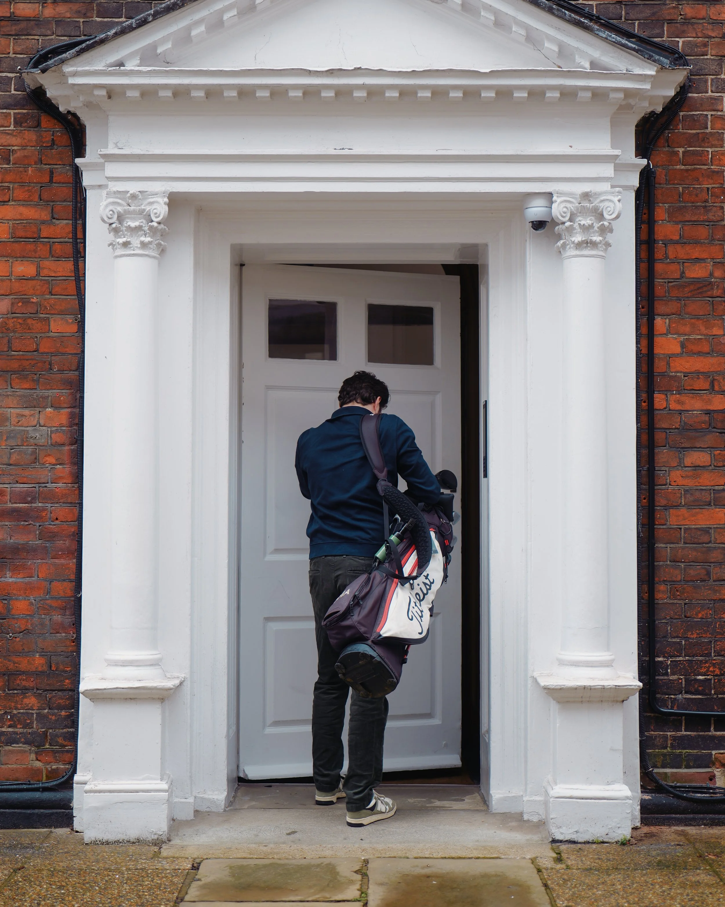 A person with a golf bag entering a house through a white door with a classical architectural frame, on a brick building.