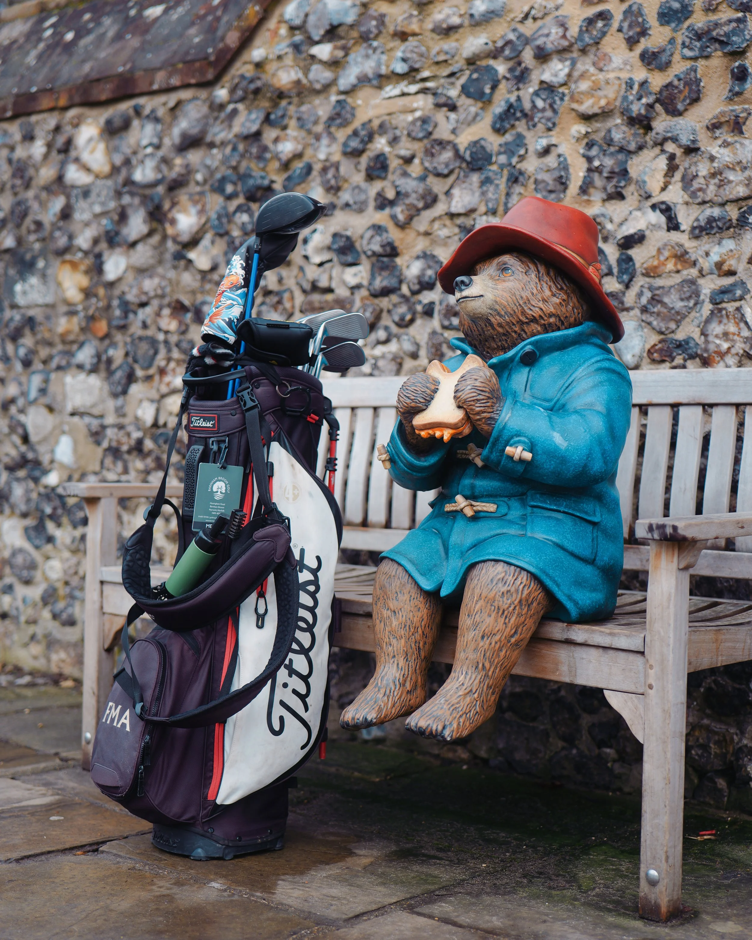A statue of a bear dressed as a golfer, wearing a red hat and blue coat, sitting on a wooden bench holding a golf club-shaped cookie or biscuit. A golf bag with clubs stands nearby against a stone wall.