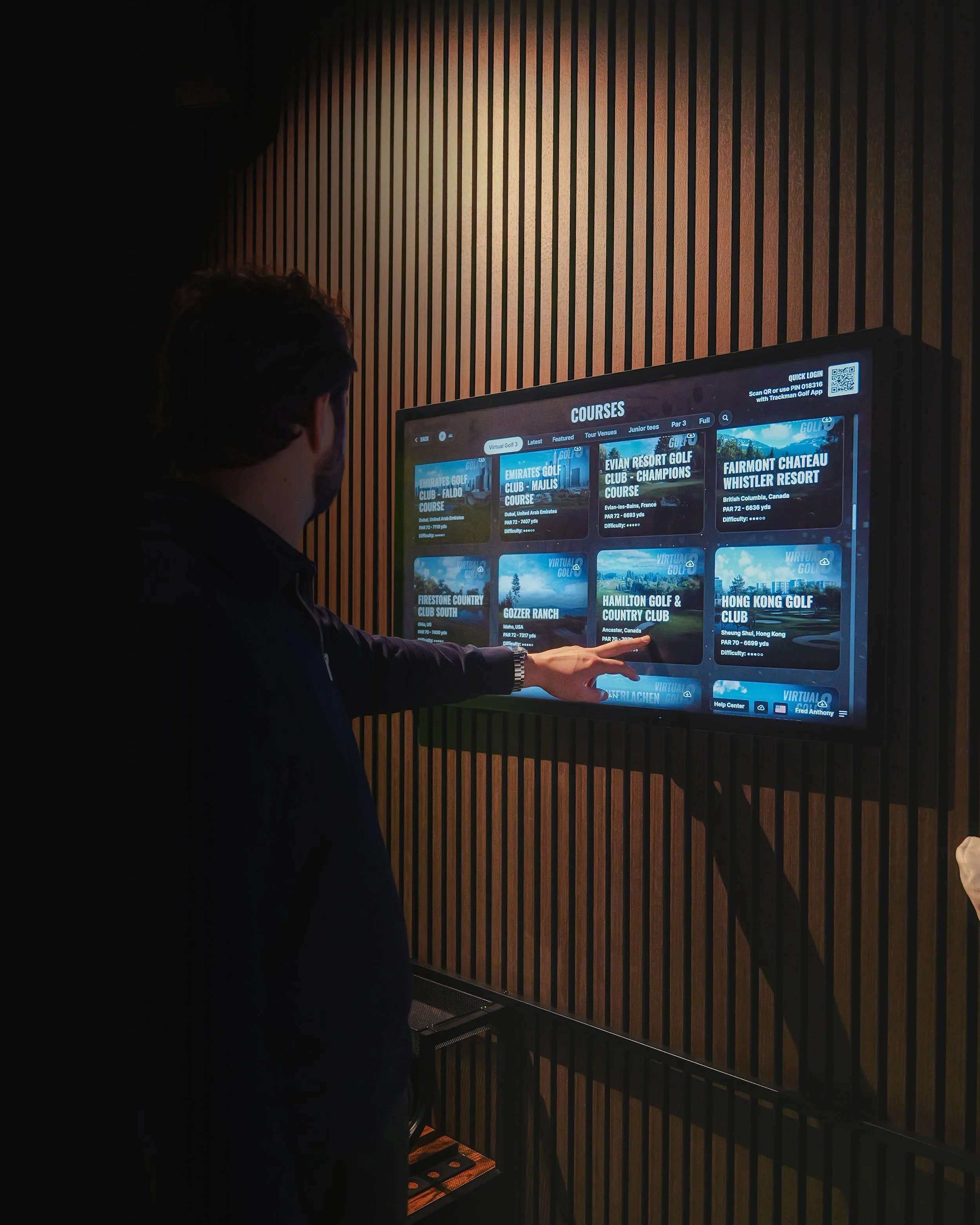 A person using a touchscreen display to browse a selection of golf courses in an indoor setting with wooden panel walls.