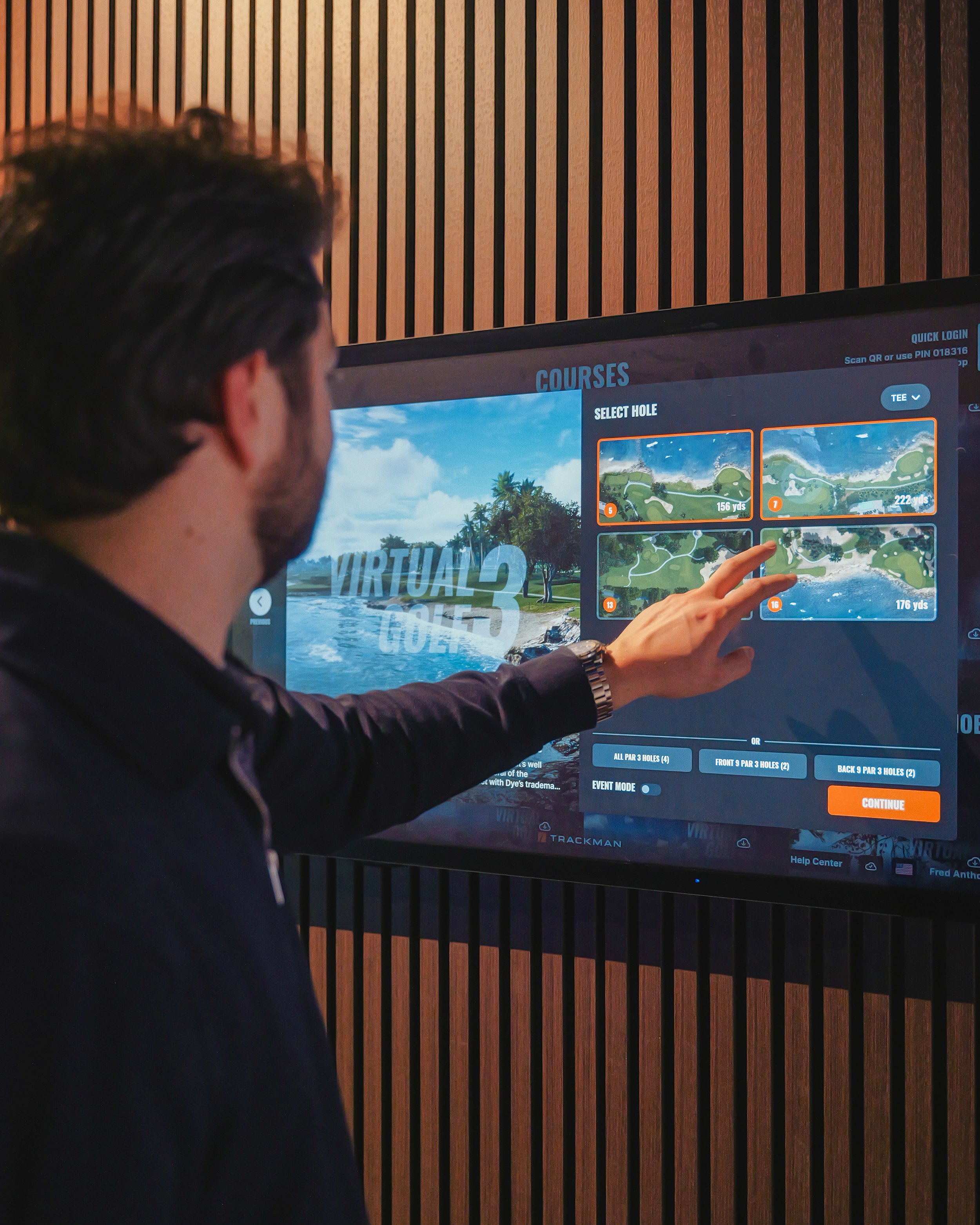 A man selecting a golf course hole on a virtual golf simulator screen in an indoor setting.