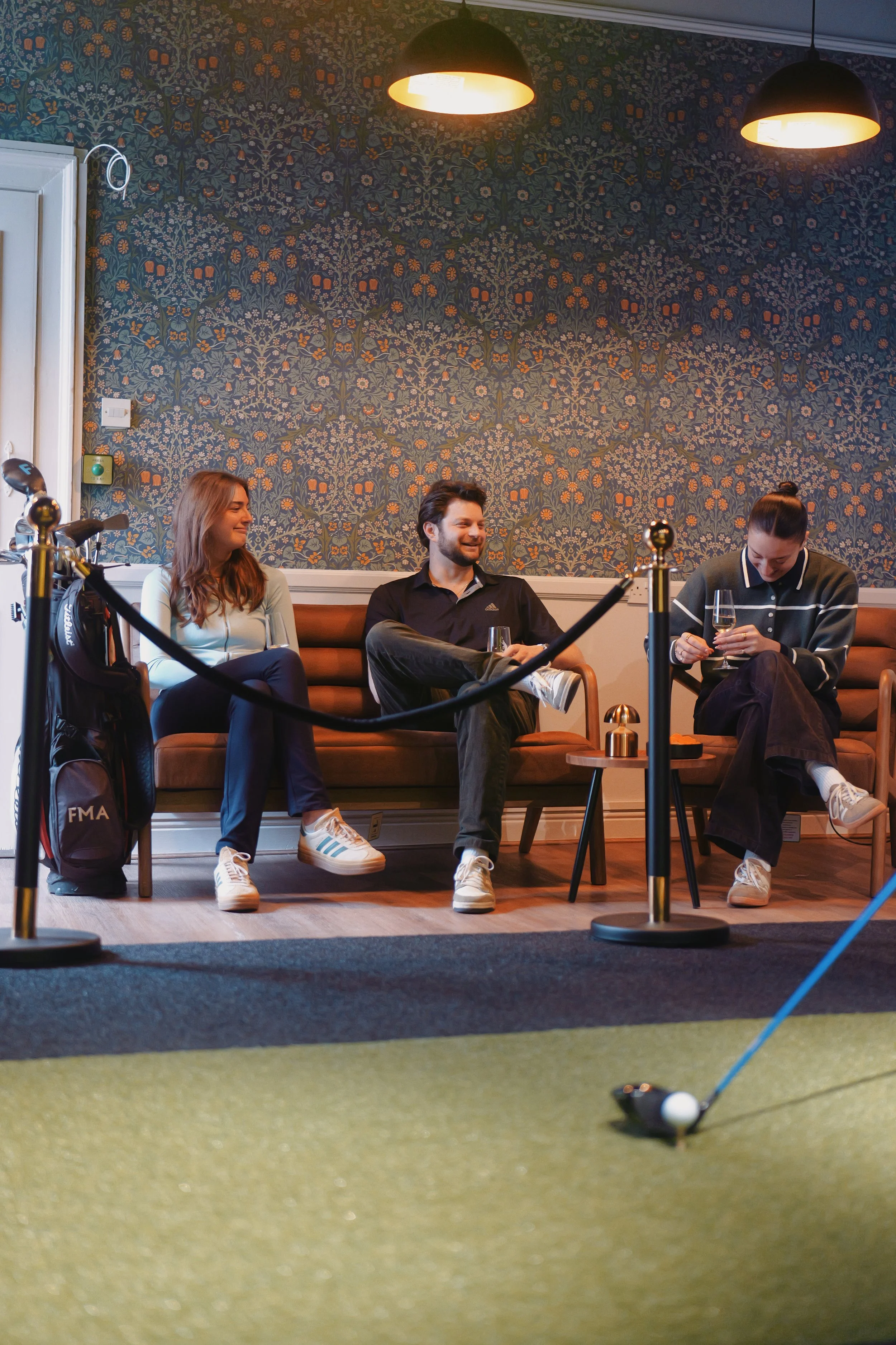 Three people sitting on a bench, two women and one man, with a golf club on the floor nearby, in front of a golf putting green inside a room, with two hanging lights overhead and a decorated blue wallpapered wall in the background.