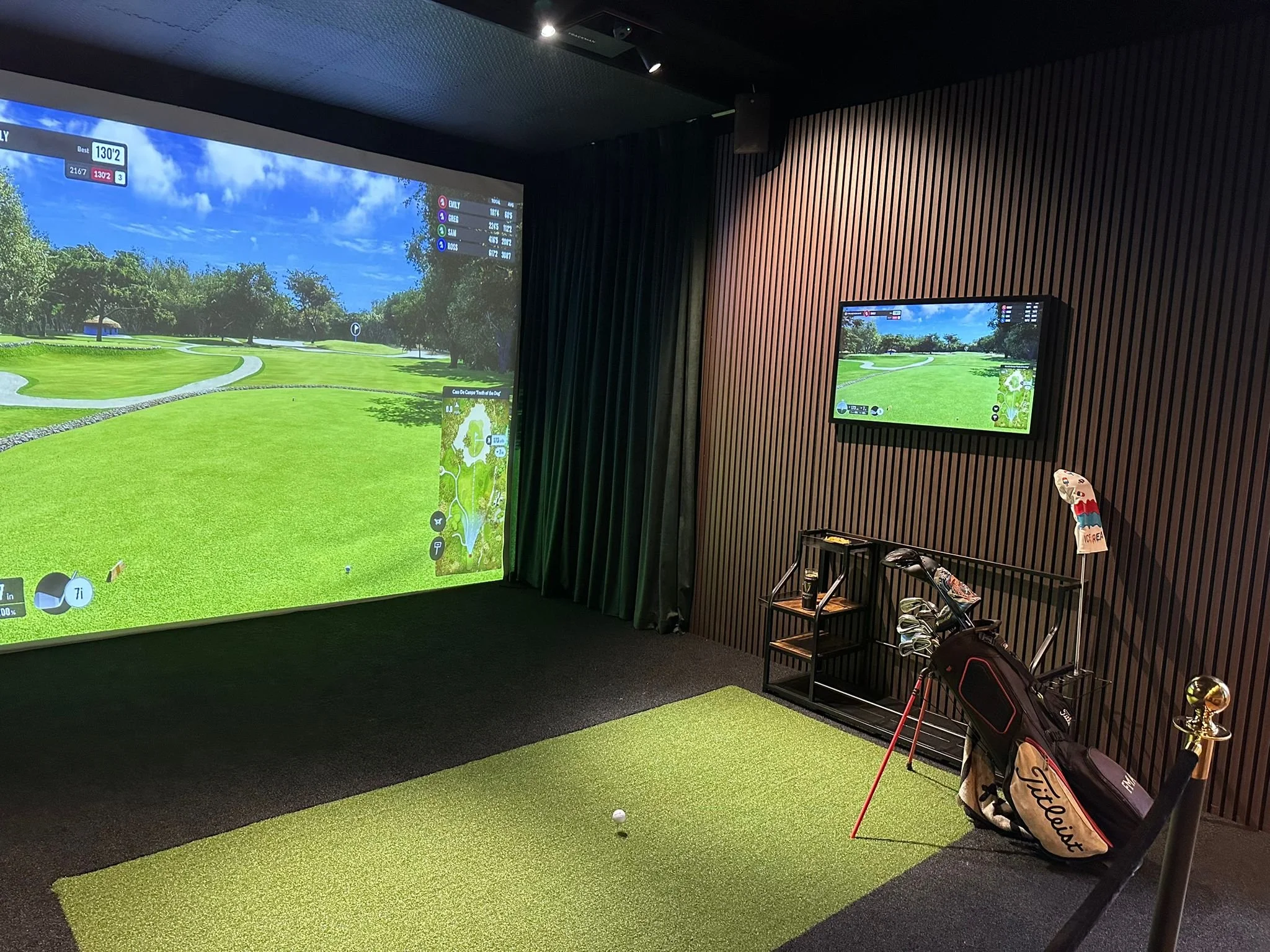 Indoor golf simulator room with a large screen showing a golf course, a smaller wall-mounted screen displaying the same golf course, a golf bag with clubs, and a golf tee on a green artificial mat.